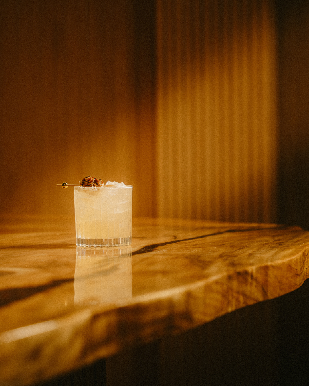 A cocktail with a skewer of a small piece of food and white garnish, served in a clear, textured glass on a wooden table.