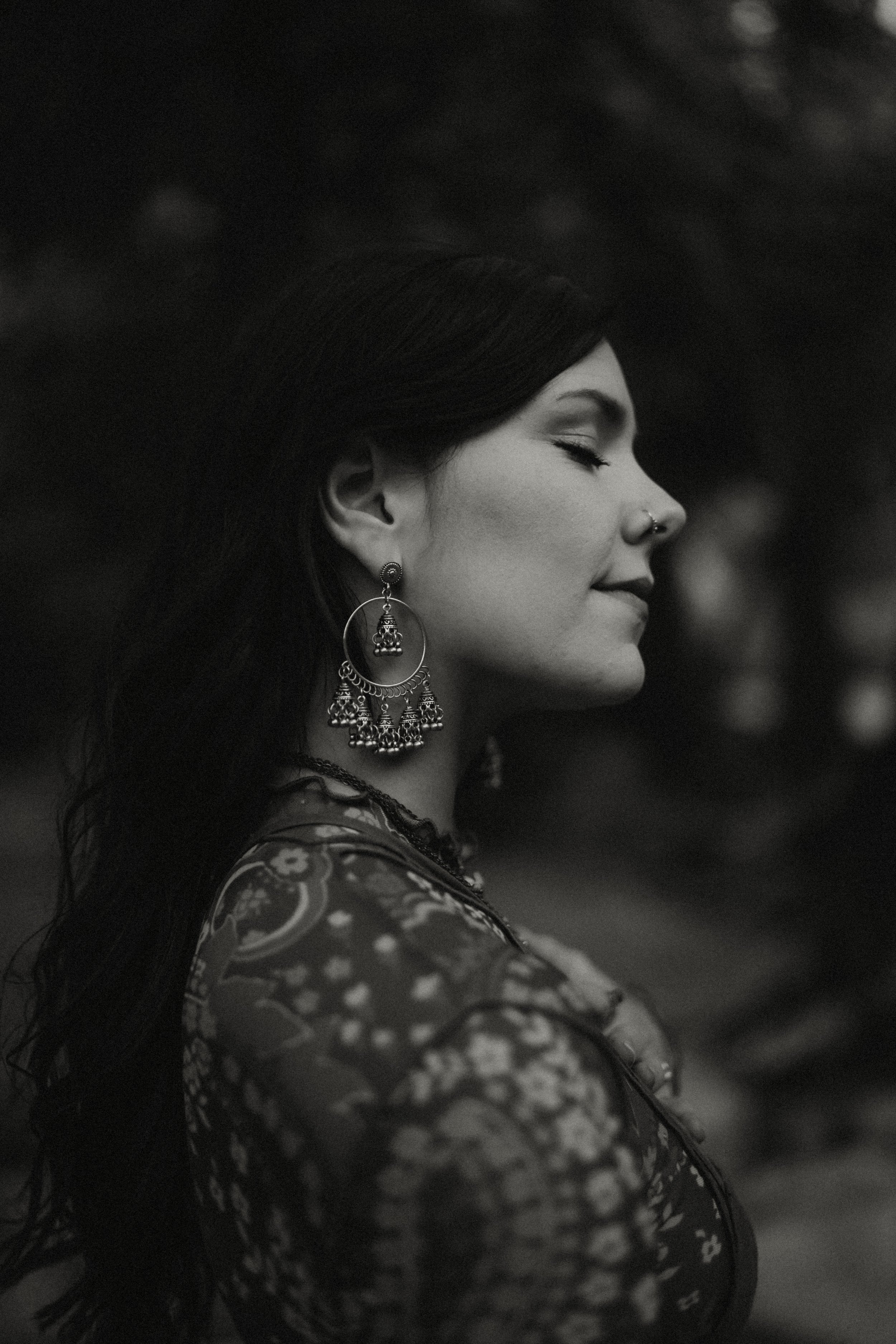 A woman with long dark hair, wearing traditional jewelry and a patterned dress, is captured in profile with her eyes closed. She has a nose ring and large earrings, and the image is in black and white.