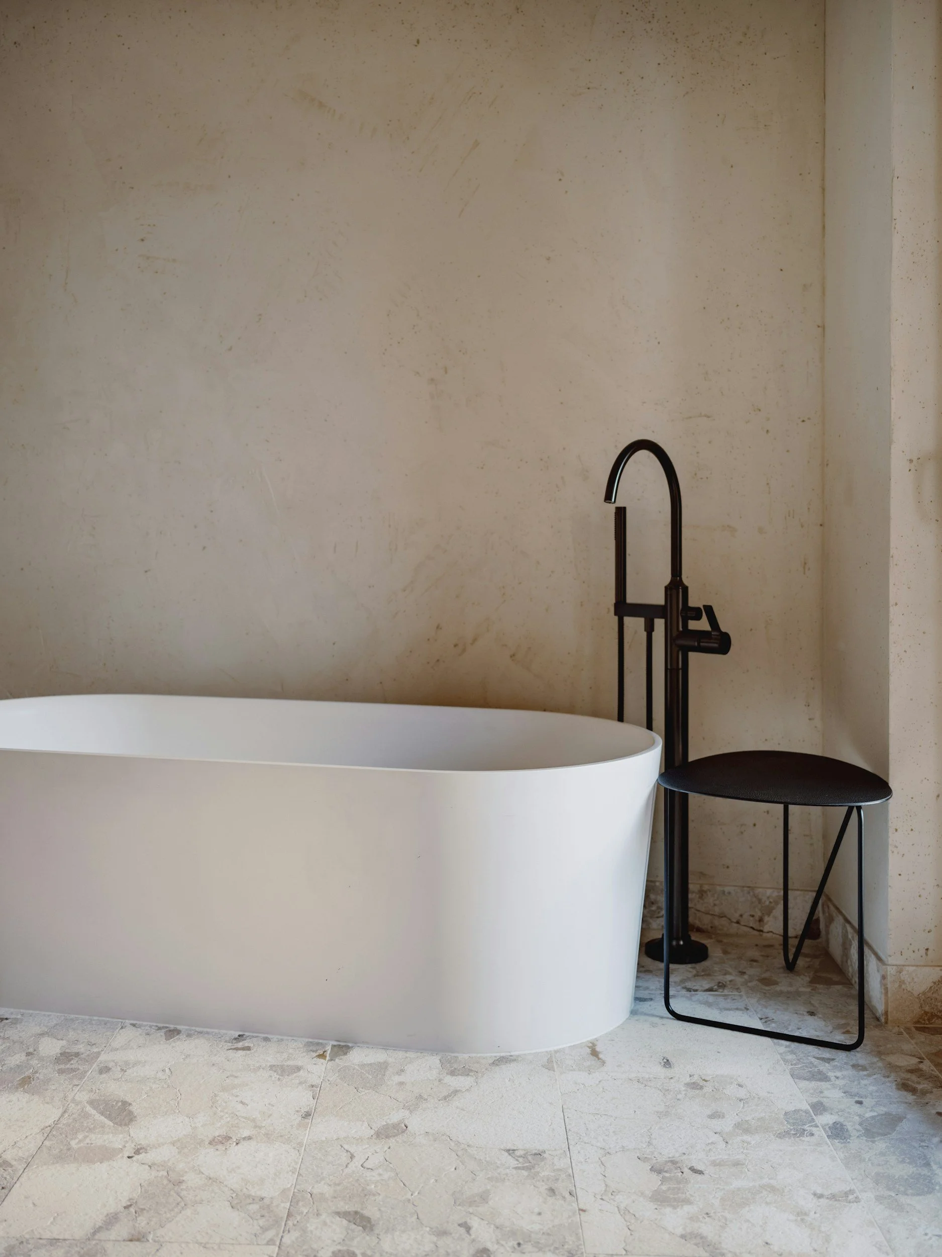 A modern bathroom with a white oval freestanding bathtub, a black floor-standing faucet, and a black chair with a round backrest. The wall behind is beige with subtle textures, and the tiled floor has a light marble pattern.
