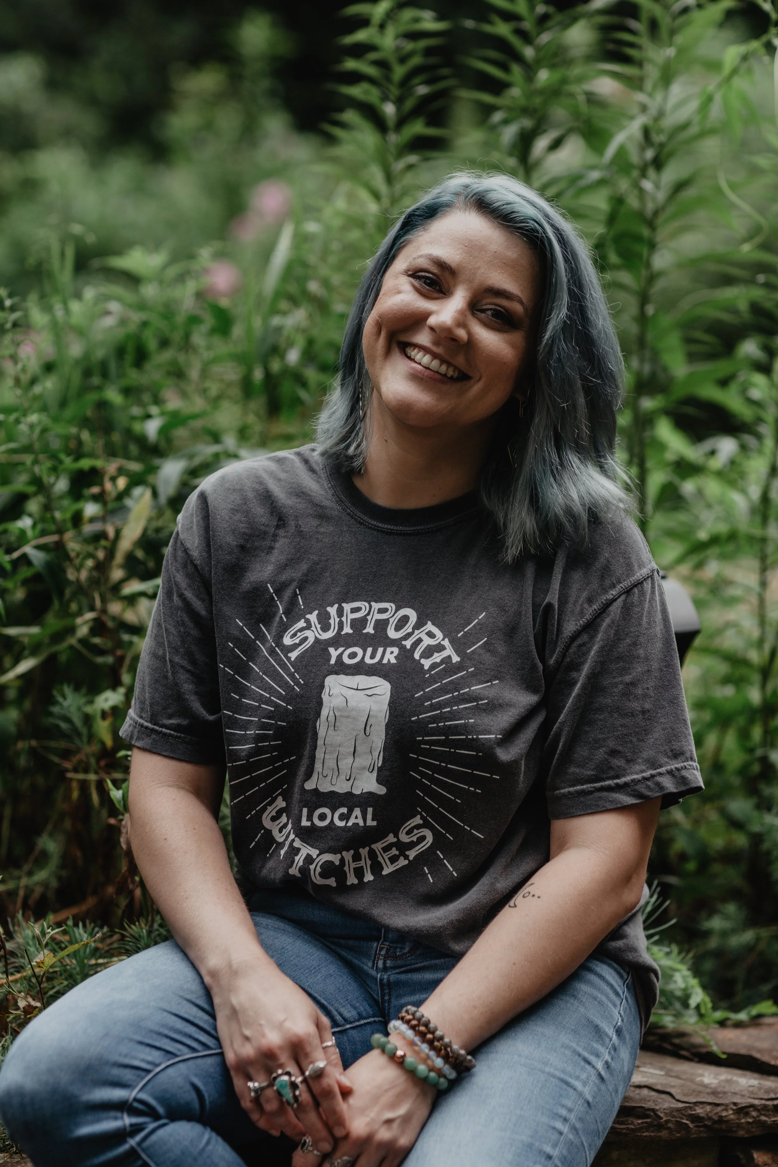 A woman with gray hair smiling while sitting on a log outdoors, wearing a dark t-shirt with a support local witches slogan and multiple bracelets on her wrist.