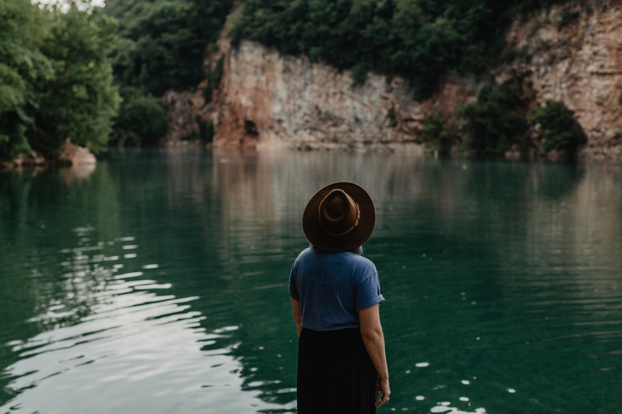 A person wearing a hat and blue shirt standing by a calm river surrounded by trees and rocky cliffs.