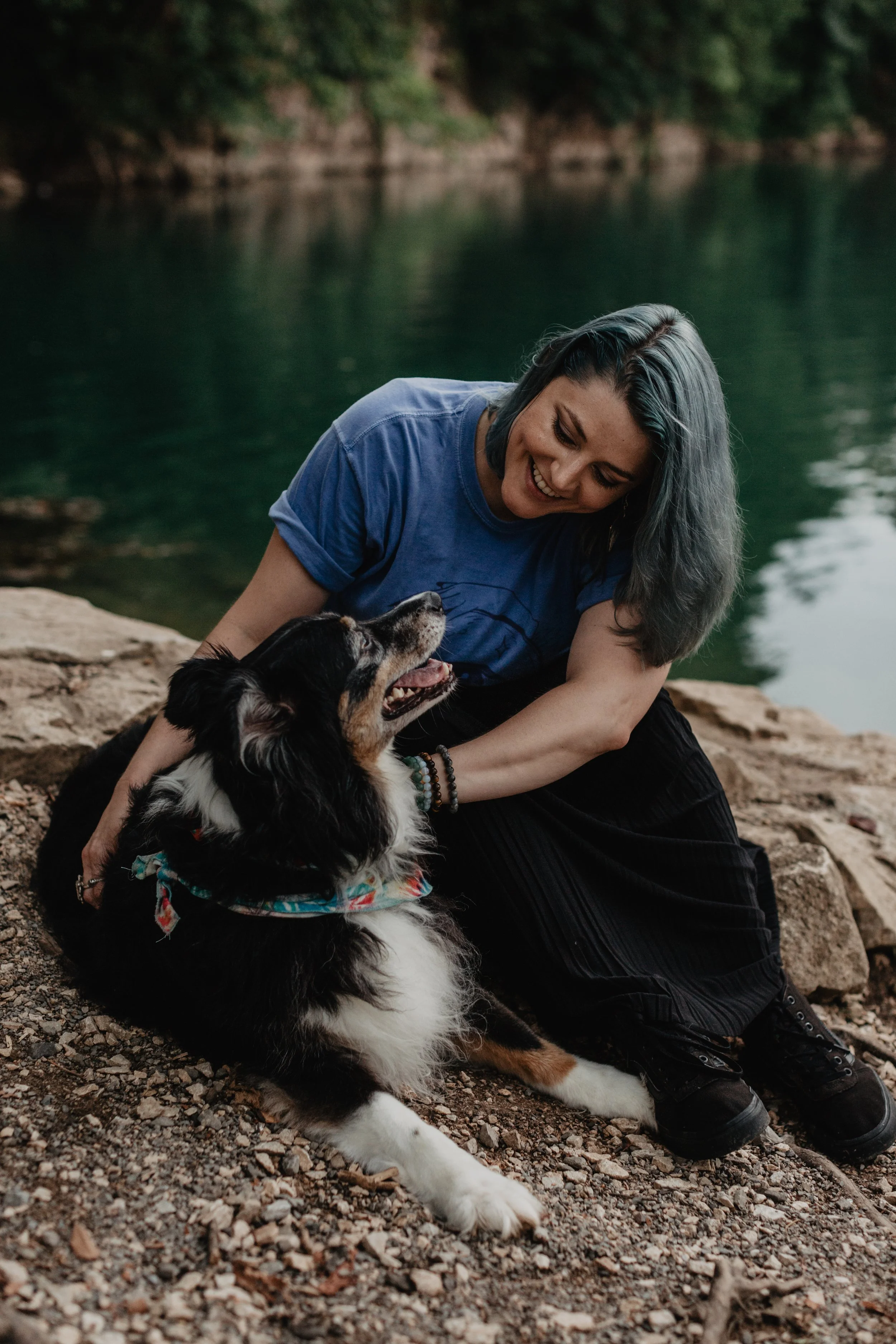 A woman with short, gray hair wearing a blue t-shirt is playing with a black, white, and brown Australian Shepherd dog near a river. The woman is smiling and leaning towards the dog, which is sitting on a rocky surface, looking up at her. The background features a calm river and lush green trees.