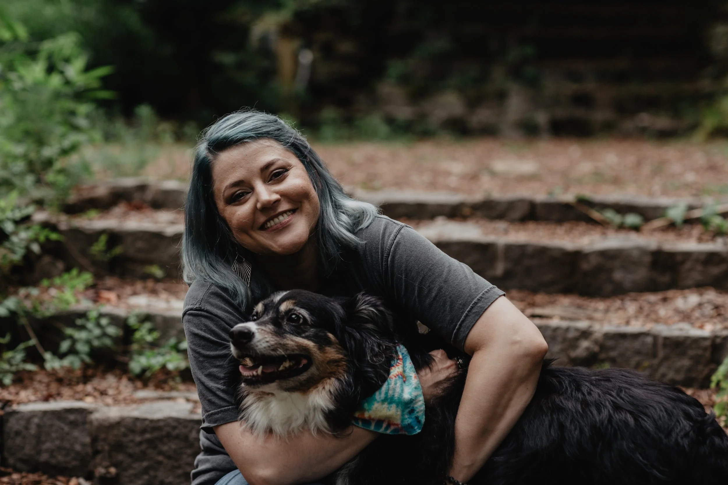 A woman with gray hair hugging her black and tan Australian Shepherd dog in a forest setting.