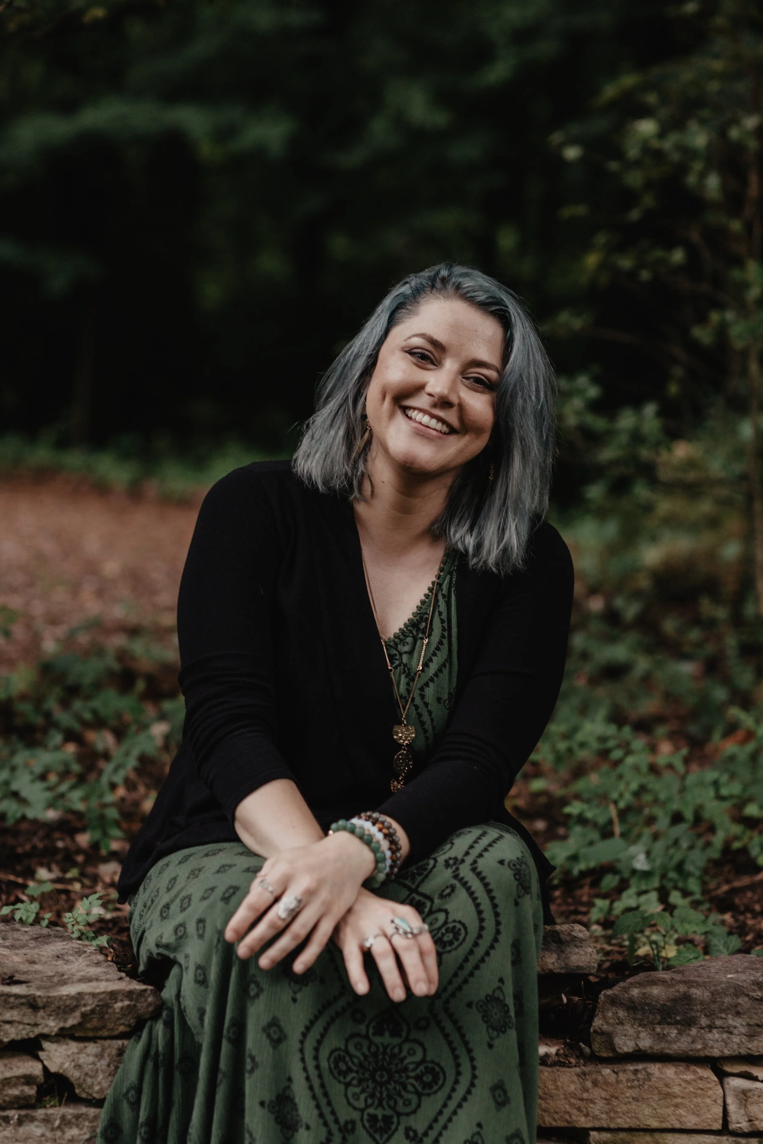 A woman with gray hair sitting outdoors on a stone structure, smiling, with a background of green trees and foliage.