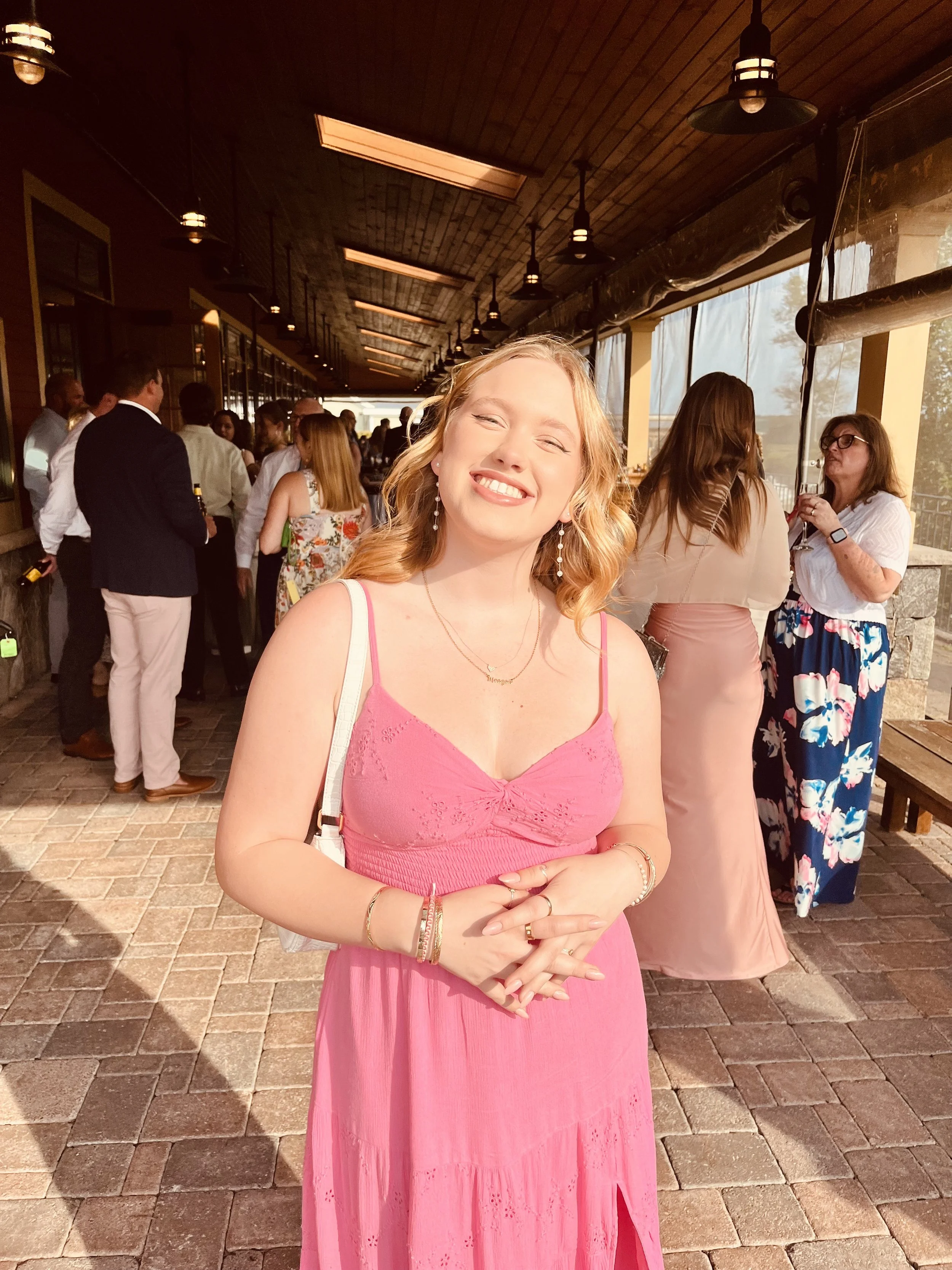 A young woman in a pink dress smiling with eyes closed, standing in a lively indoor/outdoor gathering area with several people in the background.