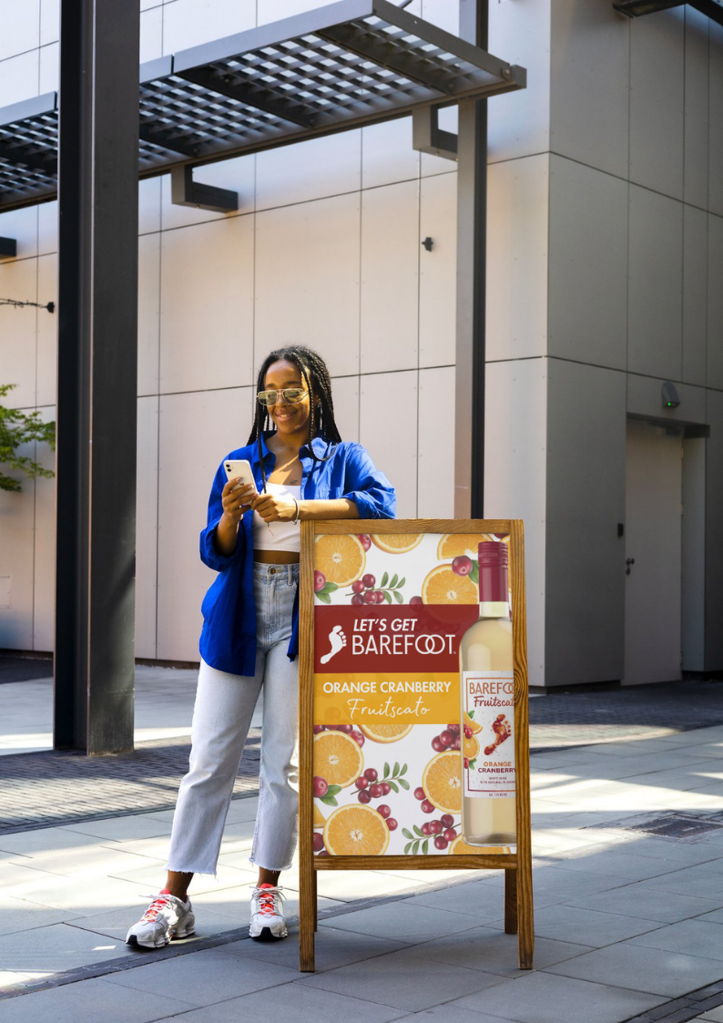 A young woman standing outdoors next to a promotional sign for Barefoot Fruitscato, a beverage with orange and cranberry flavor. She is smiling at her phone, wearing sunglasses, a blue jacket, white crop top, light blue jeans, and sneakers. The backg