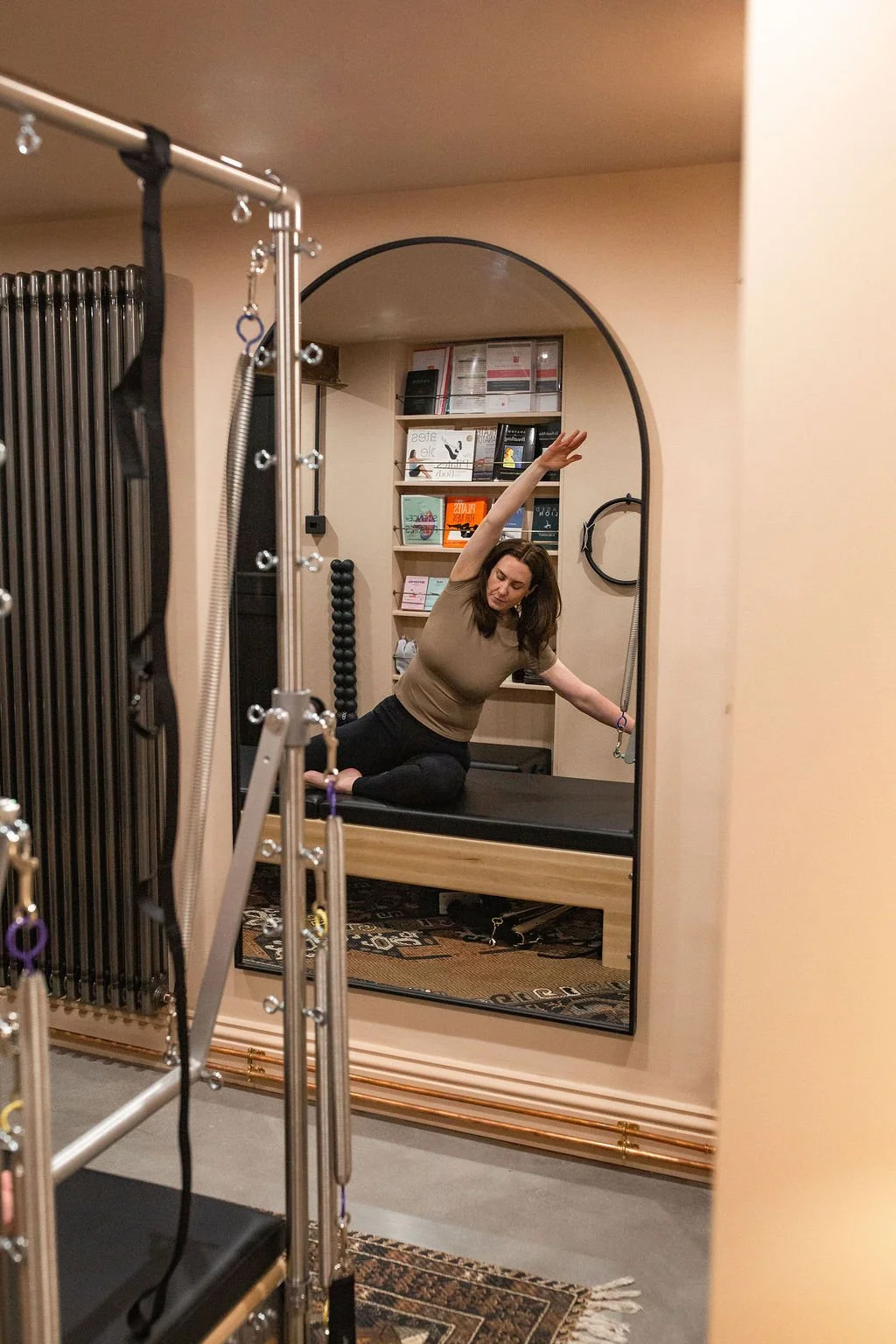 Woman practicing yoga on a Pilates reformer machine, reflected in a mirror in a studio with bookshelves and exercise equipment.