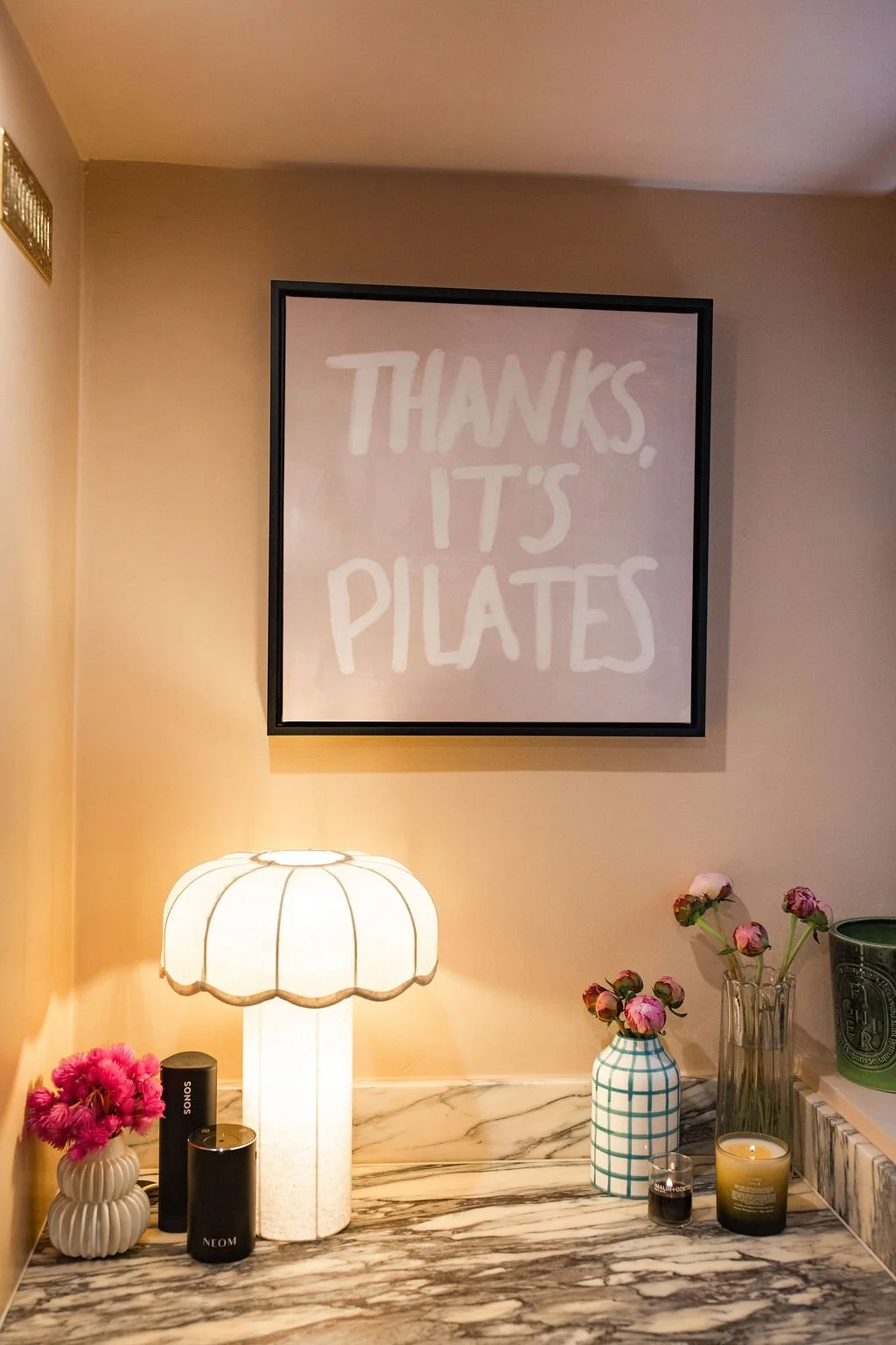 A framed sign on a beige wall reading, "Thanks, it's Pilates." There is a marble surface with a white lampshade, pink flowers in a white ribbed vase and a checkered blue vase, a candle, and various small containers.