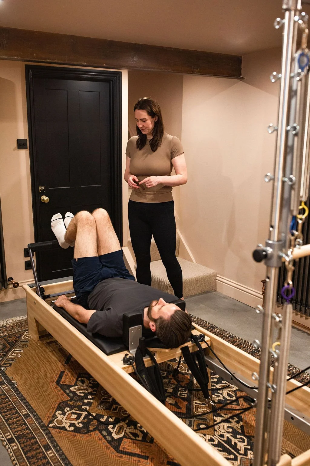 A man lying on a specialized chiropractic or physical therapy table with his knees bent, speaking with a woman standing beside him in a room with beige walls, black door, and equipment.