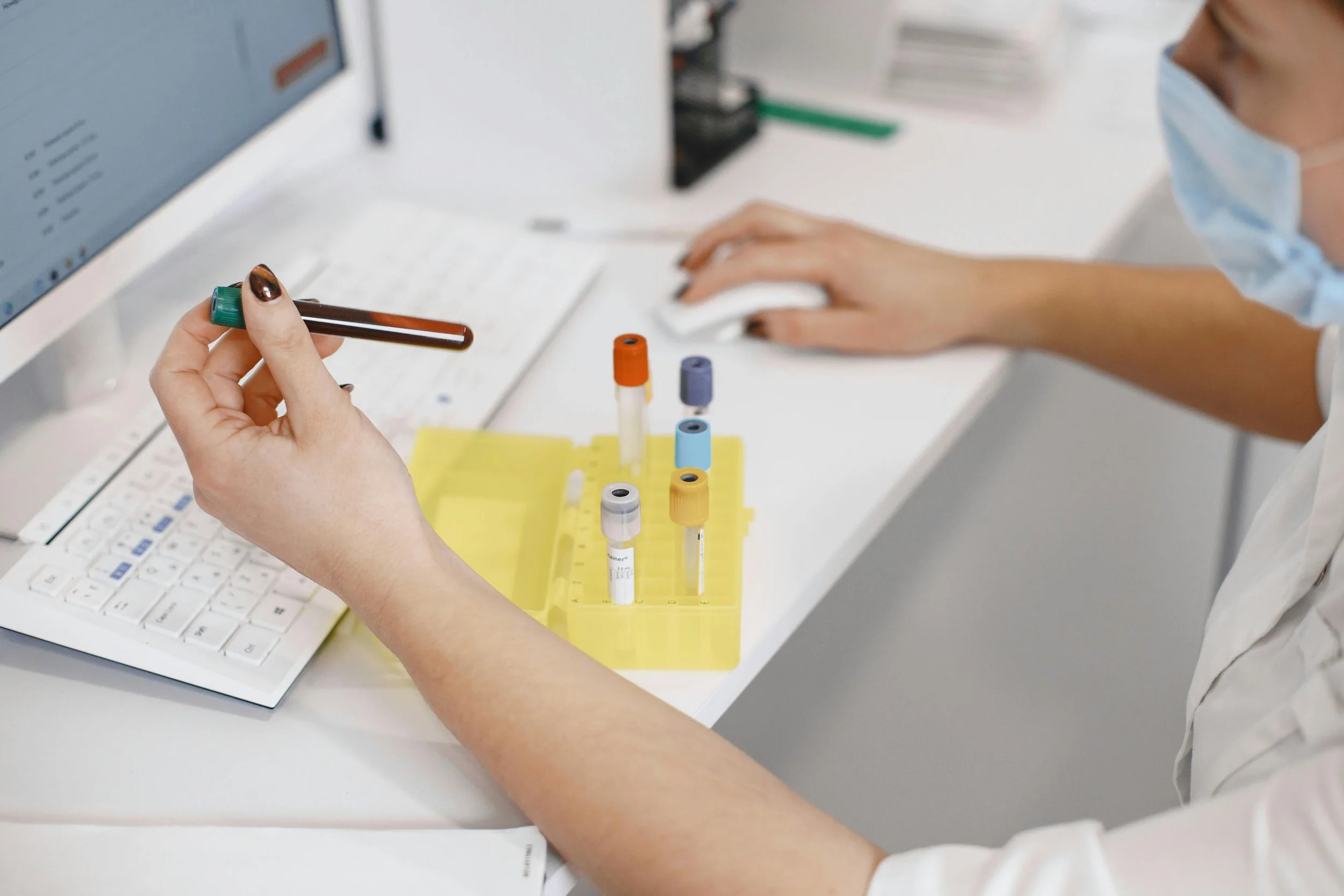 Person wearing a face mask and white clothing handling a test tube with blood in a laboratory. The test tube is held over a keyboard, with more test tubes in a yellow rack nearby.