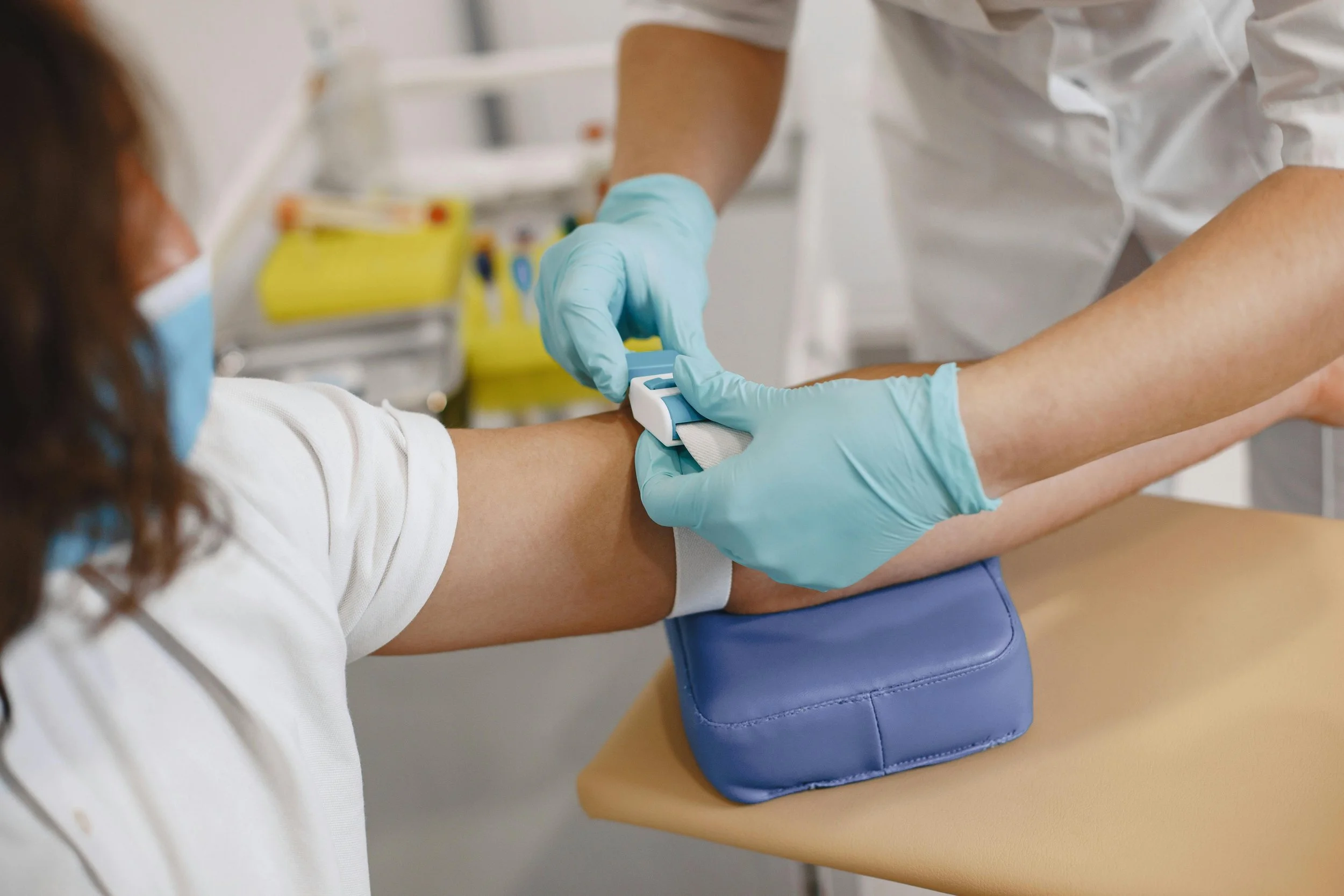 A healthcare professional wearing blue gloves is preparing to draw blood from a patient's arm using a tourniquet and a needle in a clinical setting.