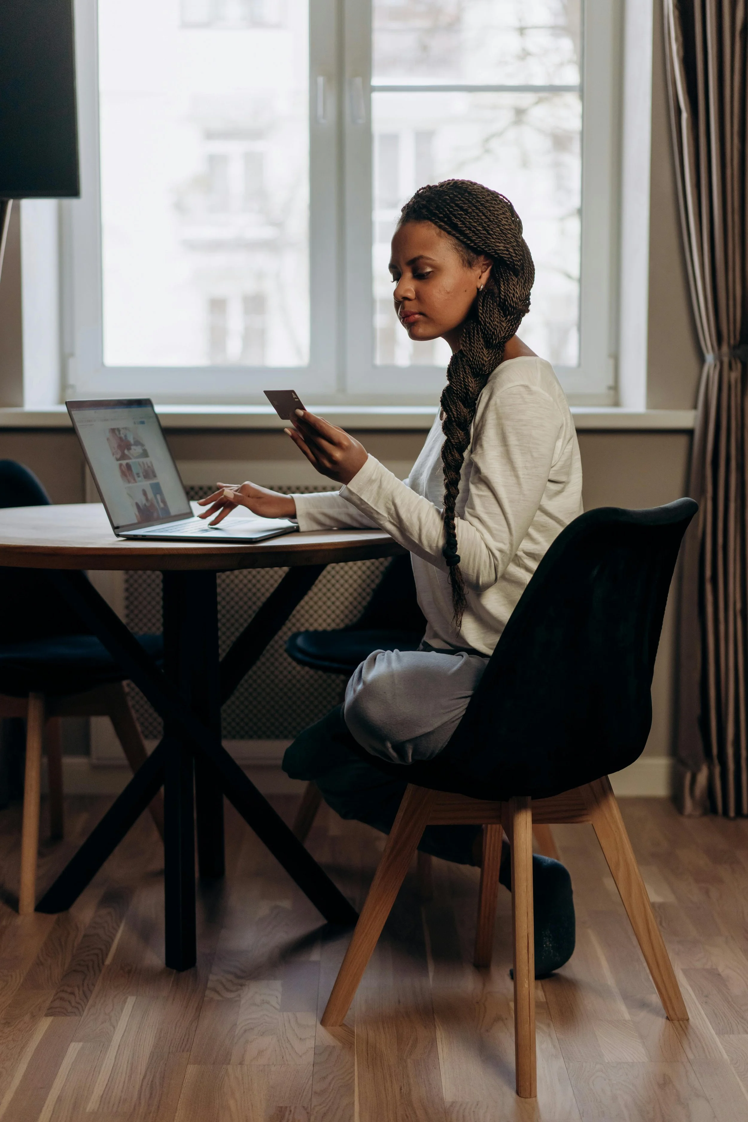 A woman with braided hair sitting at a wooden table in a room, using a laptop and holding a credit card, with a window and curtains in the background.