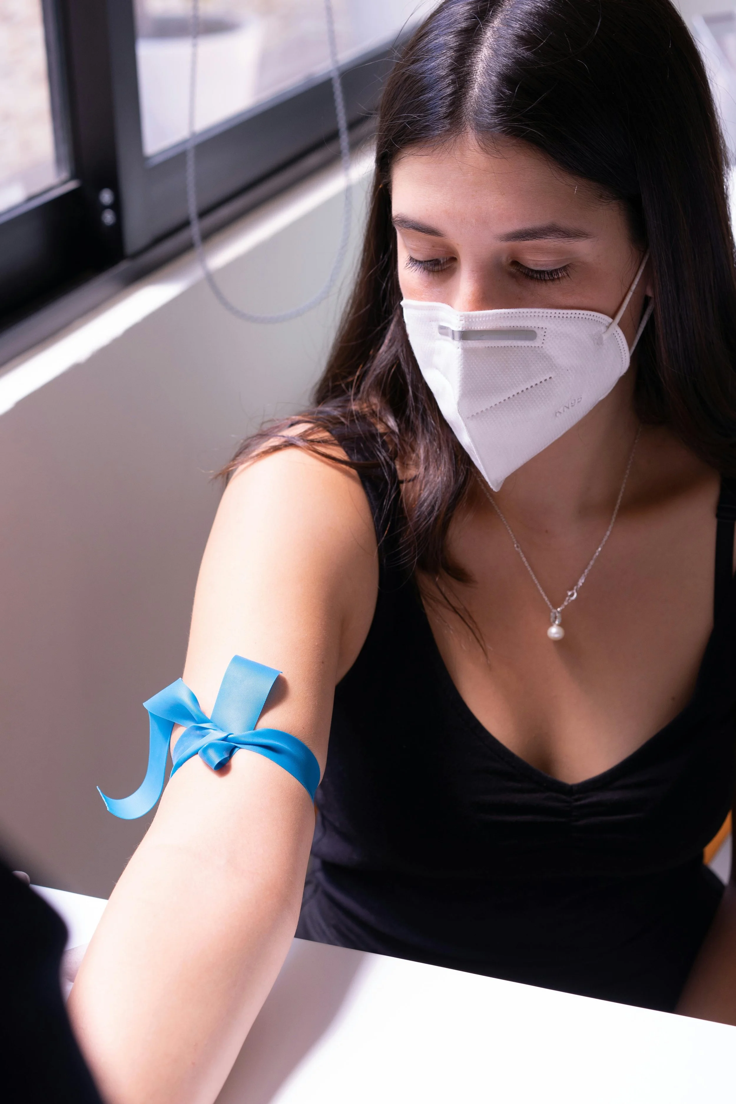 A woman wearing a white face mask and a black sleeveless top is donating blood. A blue ribbon is tied around her upper arm, and she is sitting indoor near a window.