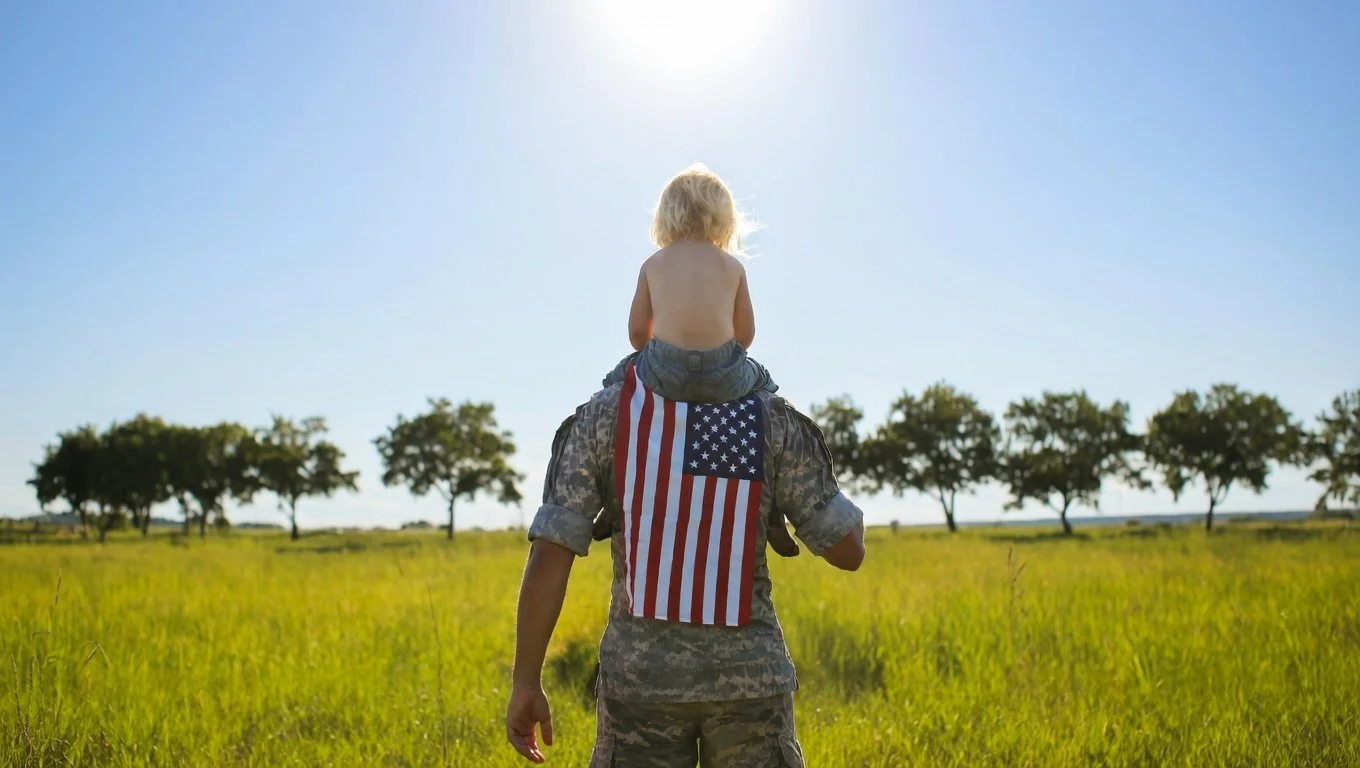 A soldier carrying a young child on their shoulder in a grassy field with trees under a clear blue sky.
