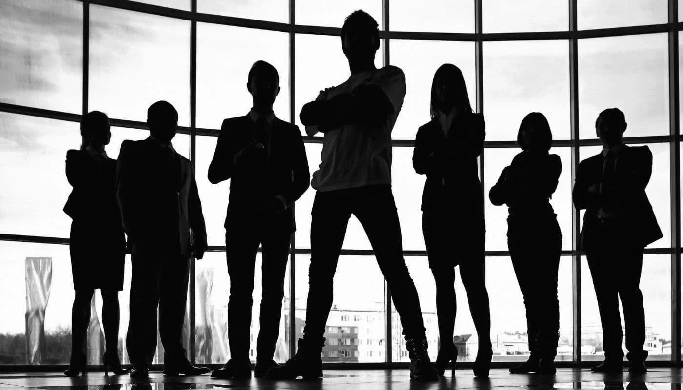 Silhouette de siete personas de negocios en una sala de reuniones con grandes ventanas que dejan ver un paisaje urbano al fondo.