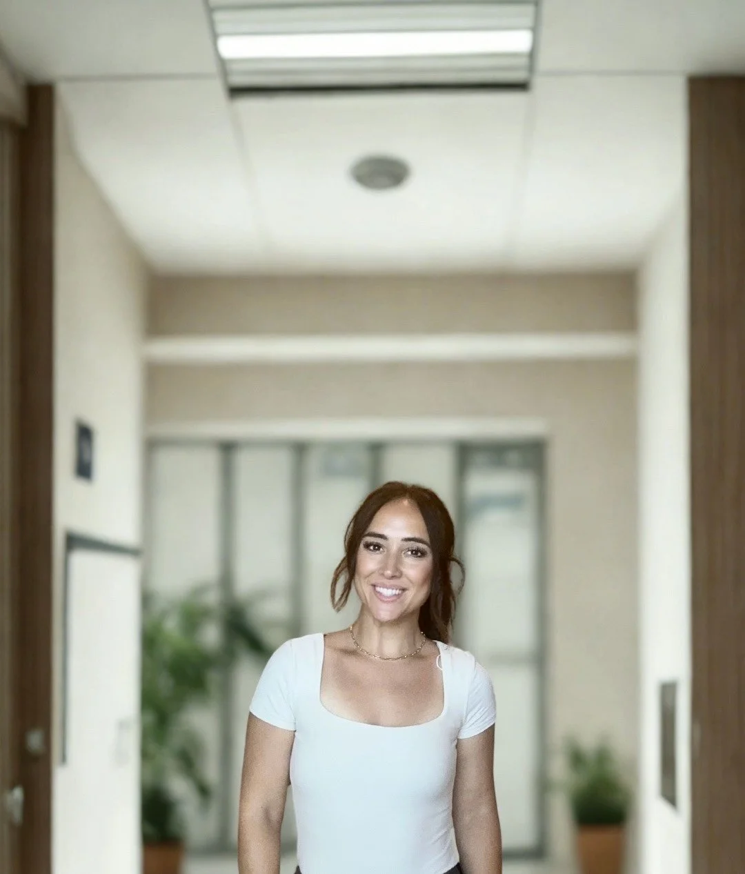 Smiling woman with brown hair in a white top standing in an indoor hallway with glass doors and potted plants in the background.