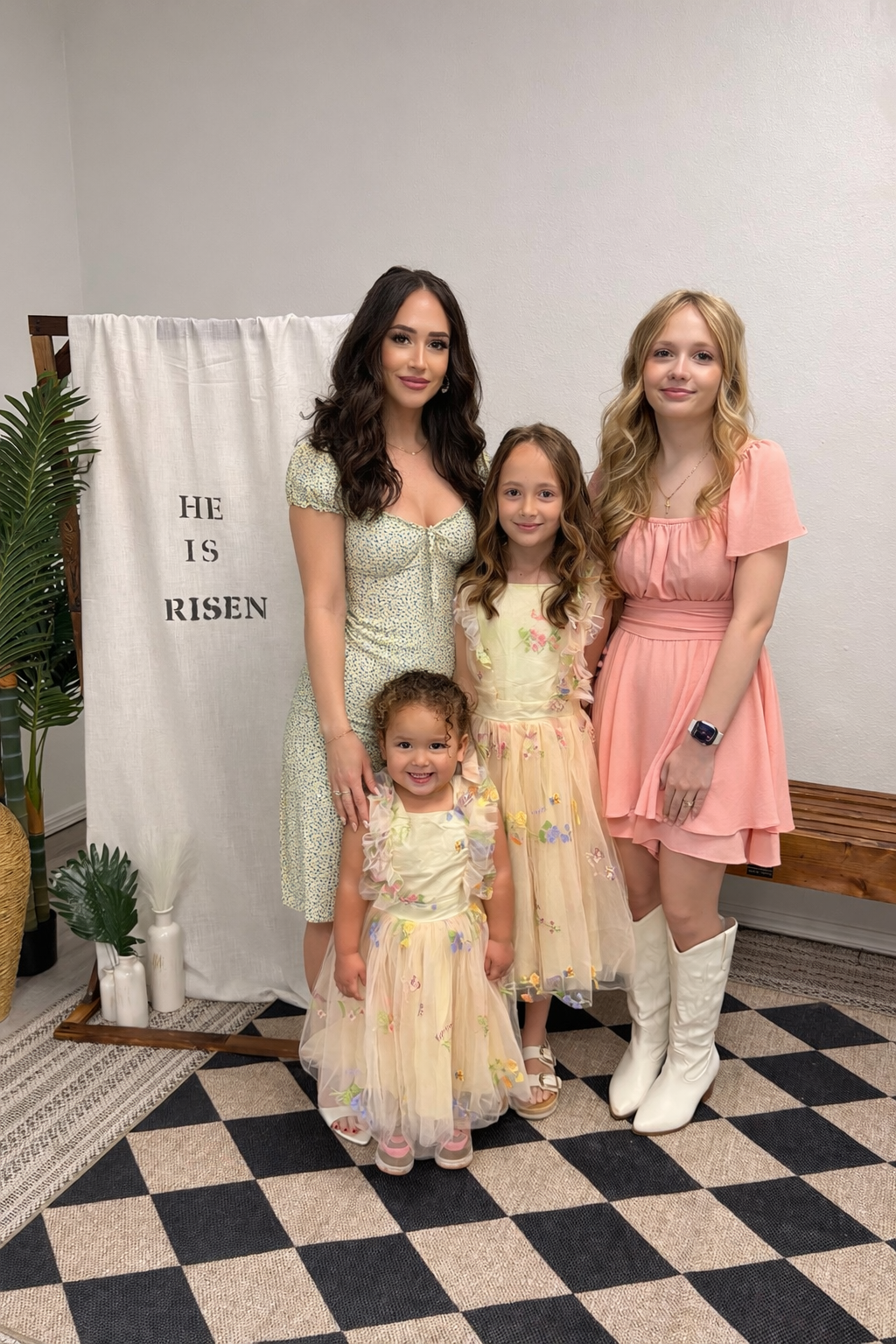 A woman with dark hair and two young girls with light hair and two young girls with curly hair, dressed in pastel-colored dresses, standing together in front of a white backdrop with the words 'He is Risen', indoors with a plant and bench nearby.
