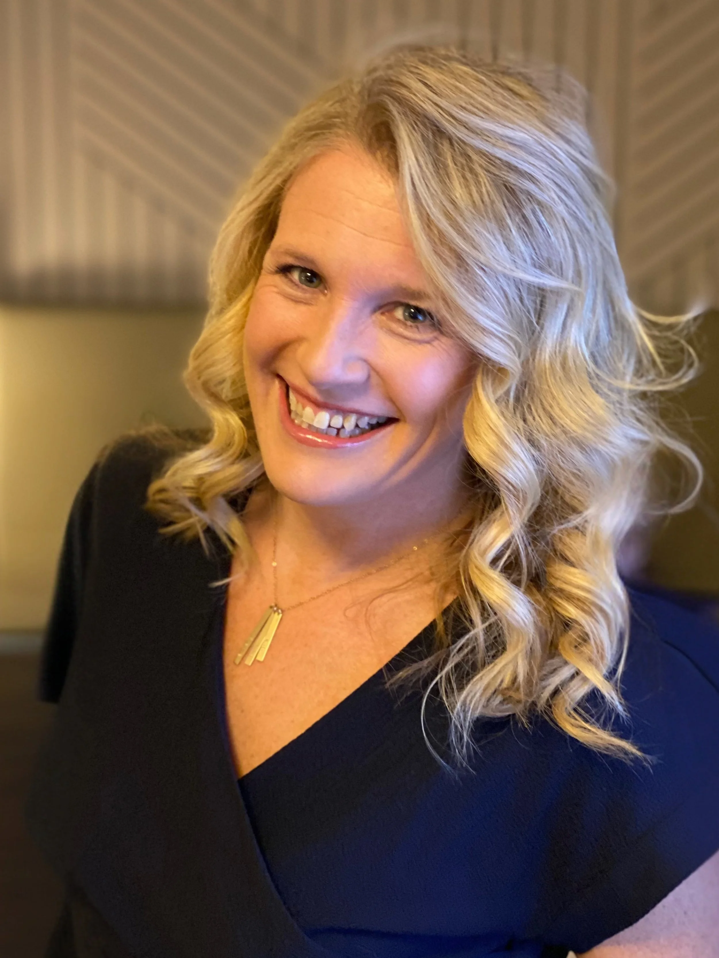 A woman with blonde, curly hair, smiling, wearing a black top and a gold necklace, in an indoor setting.