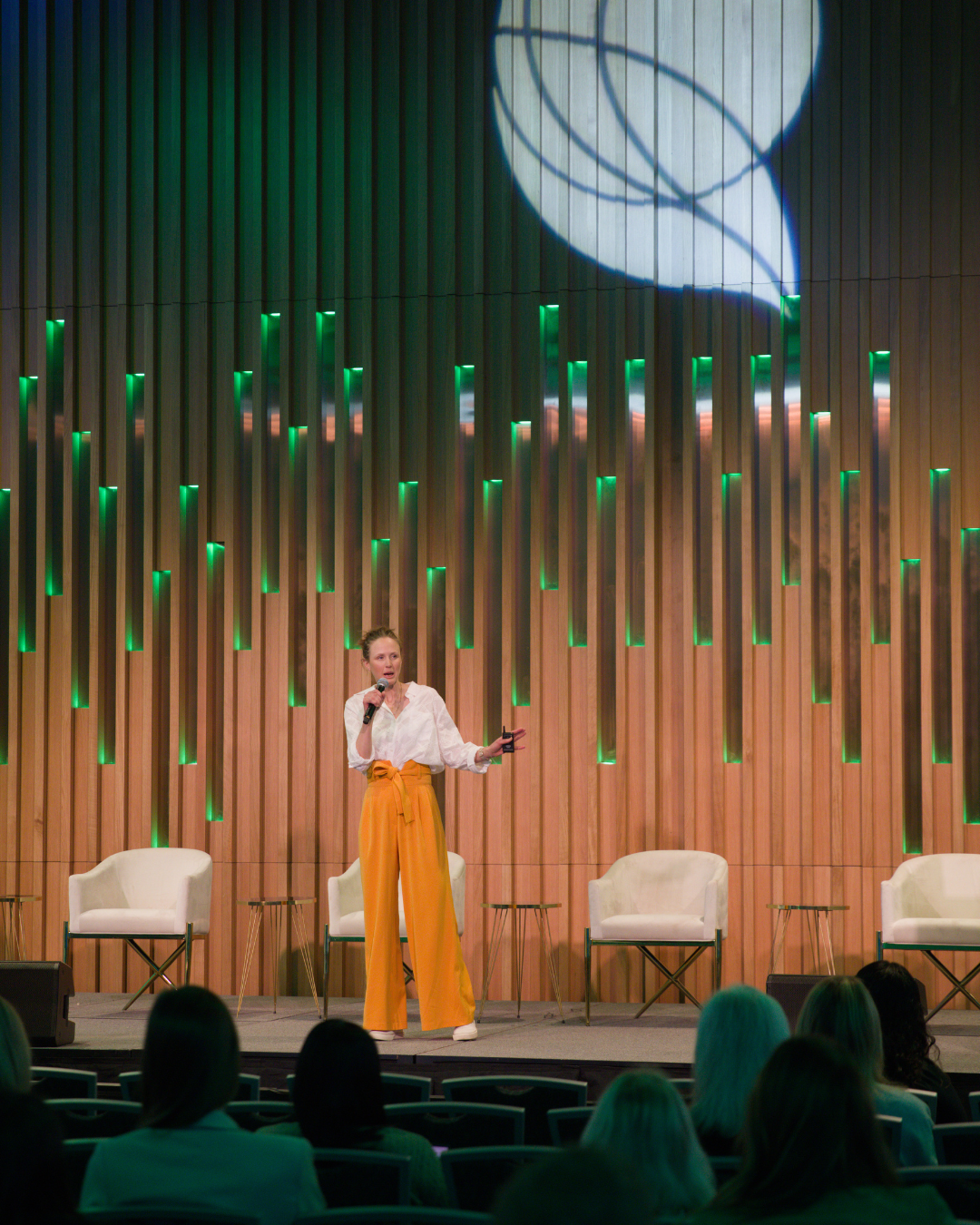 A woman speaking on stage at a conference with a microphone, wearing orange pants and a white blouse, in front of a wooden panel backdrop with green lighting accents.