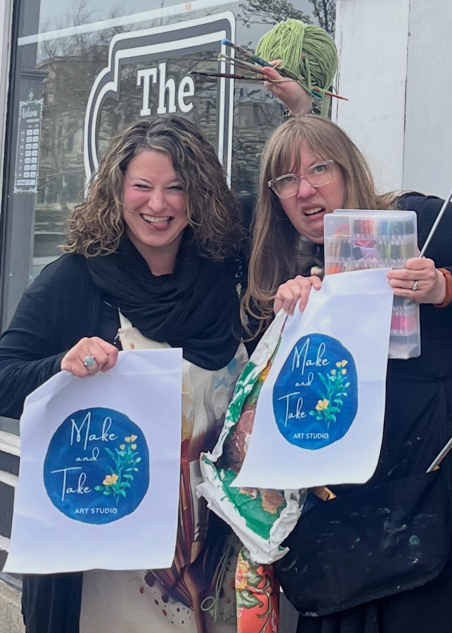 Two women smiling and holding tote bags that say "Make and Take Art Studio" outside a building with a glass door.