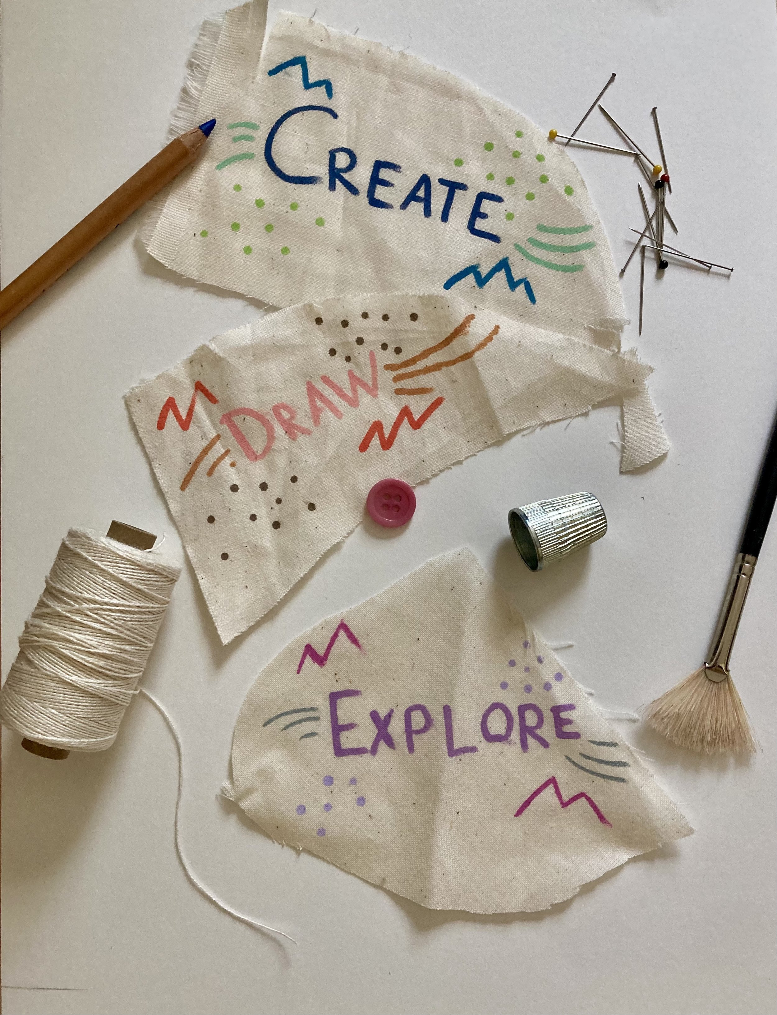 Pieces of fabric with colorful handwritten words 'Create', 'Draw', and 'Explore' surrounded by doodles, along with sewing supplies including thread, pins, a paintbrush, and a small metallic container.