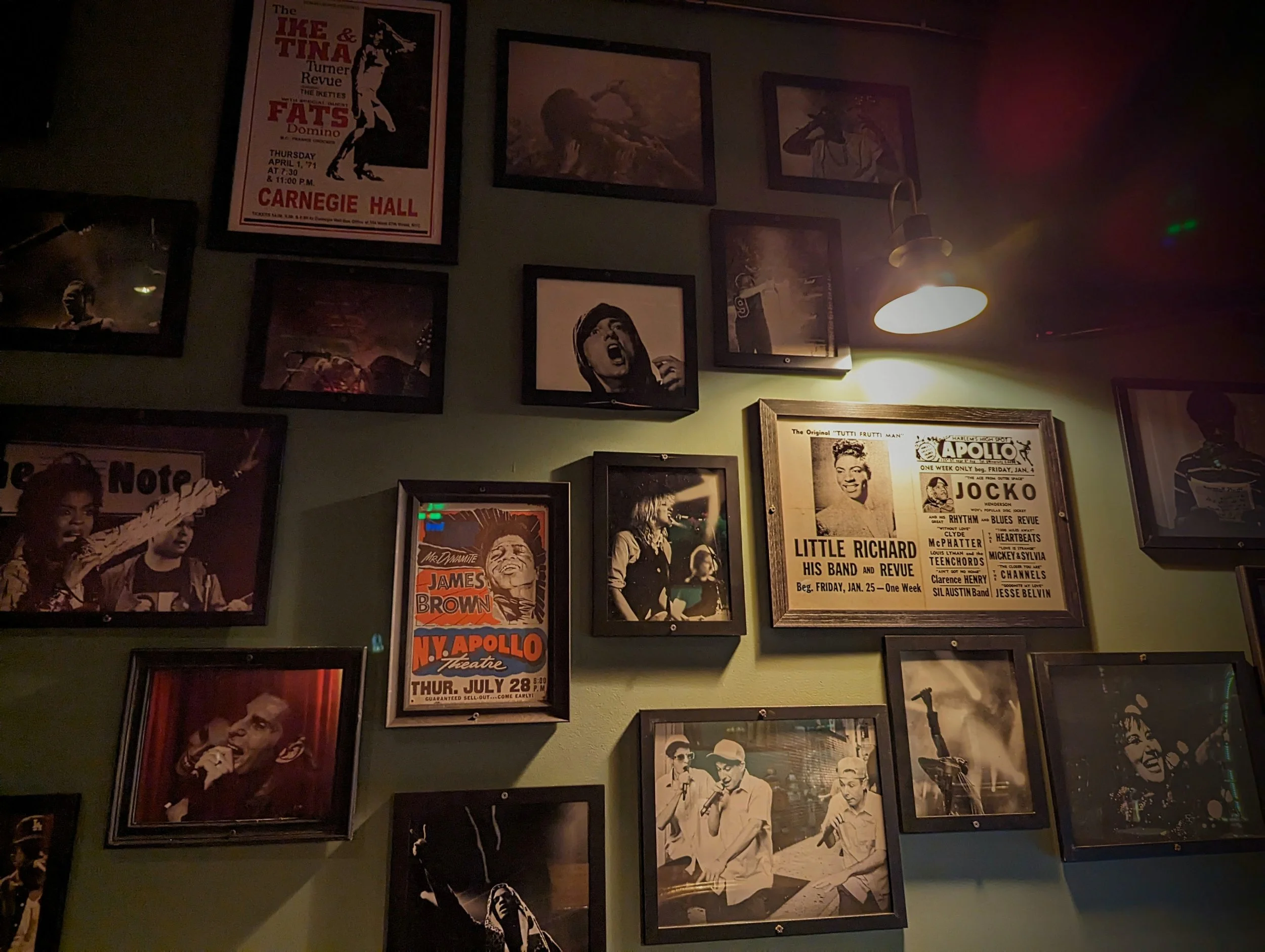 Wall with framed vintage posters, black-and-white photographs, and artwork, illuminated by a wall-mounted light fixture, in a dimly lit setting.