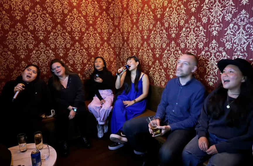 Six people singing and laughing at a karaoke night in a room with red and gold ornate wallpaper, some holding microphones and drinks.