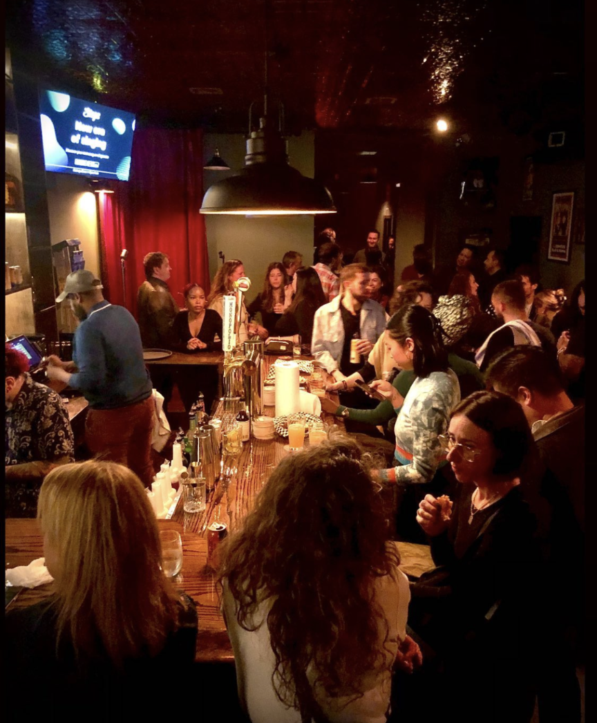 A lively bar scene filled with people socializing and enjoying drinks. The bar counter has various bottles and glasses, a bartender serving drinks, and a large hanging light fixture overhead. The atmosphere is warm and bustling with conversations.