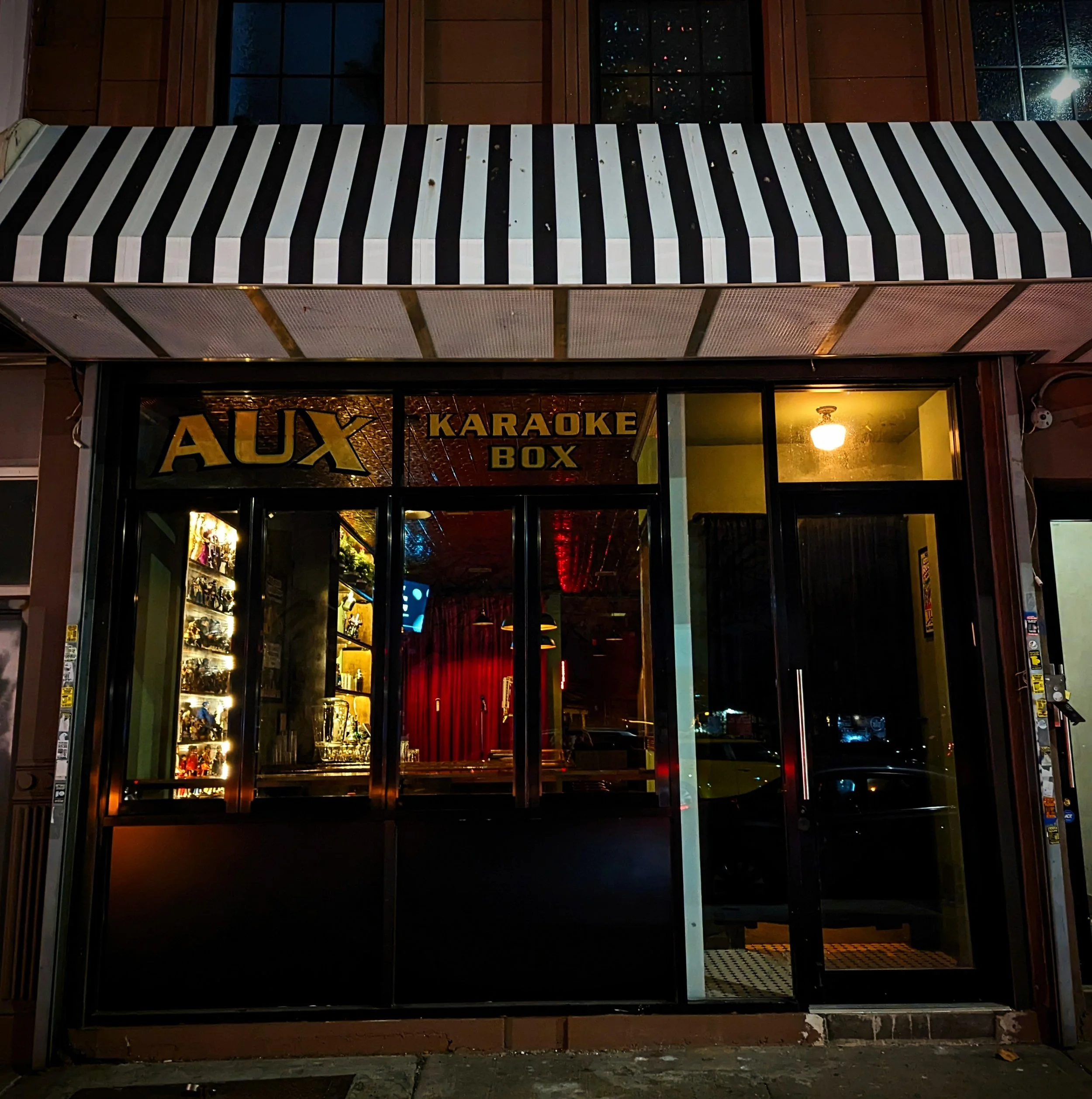  storefront with black and white striped awning, illuminated sign reading 'AUX Karaoke Box', and interior with red curtains and decorative lighting visible through glass windows.