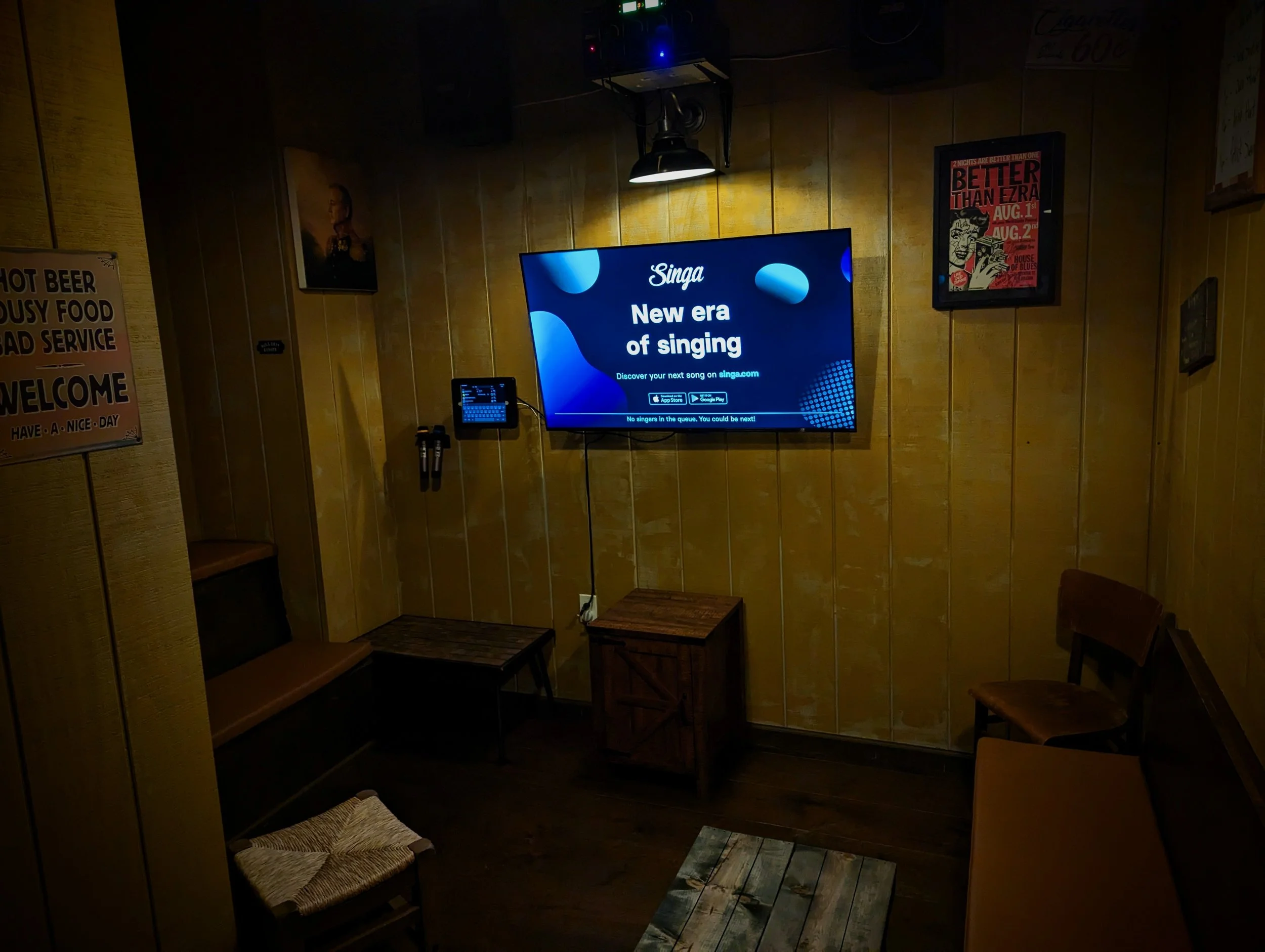 A cozy room with wooden walls, a wall-mounted television displaying an advertisement for 'Singa' with the text 'New era of singing', a small bench, a chair, and framed posters on the wall.