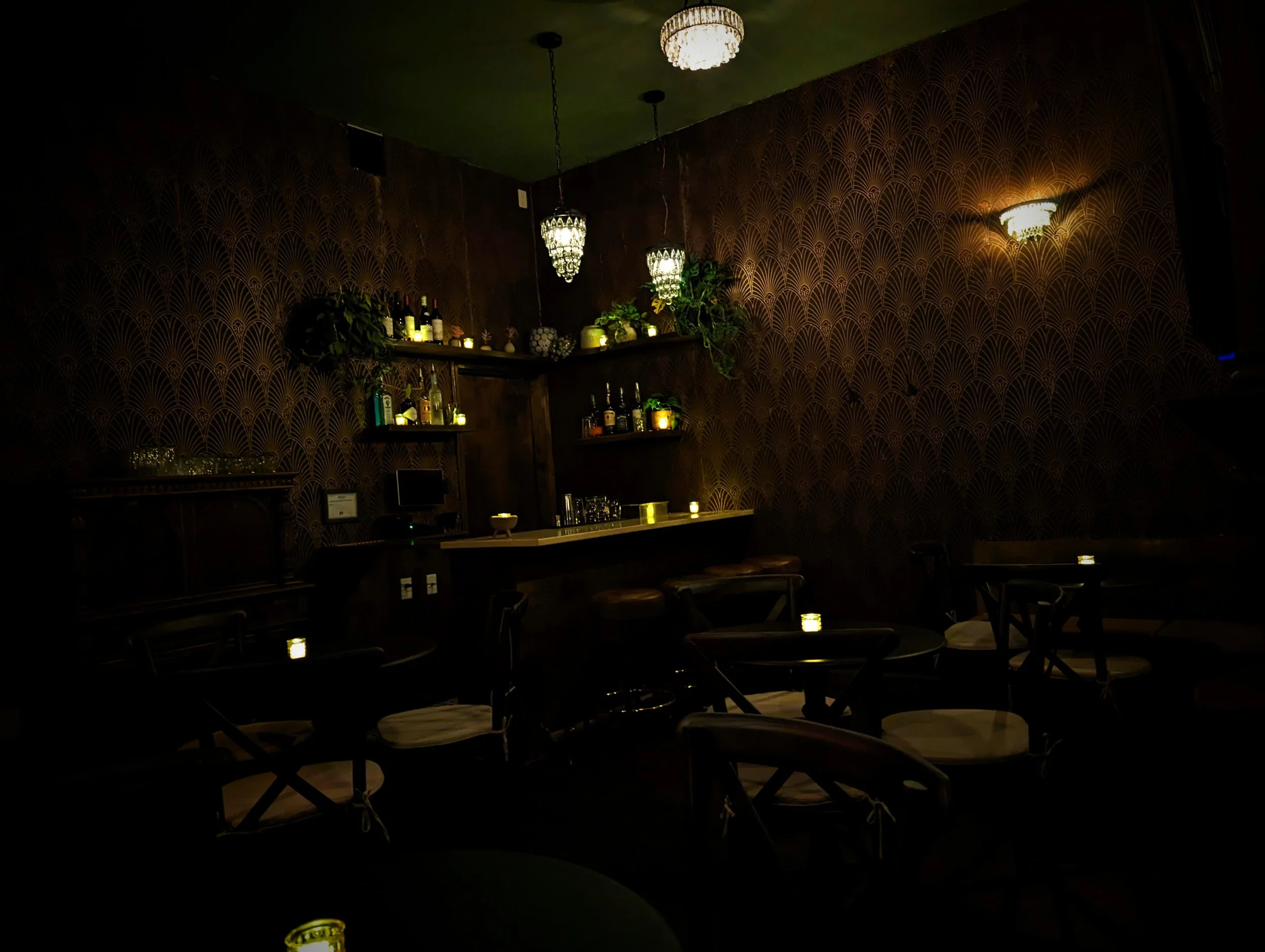 A dimly lit bar with dark wood furniture, patterned wallpaper, shelves with bottles and plants, and small candle-like lights on tables.
