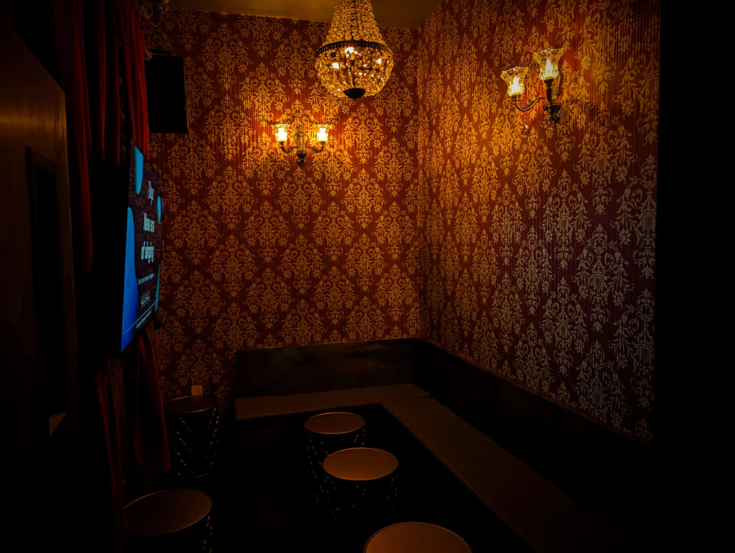 A dimly lit room with ornate gold-patterned wallpaper, a chandelier hanging from the ceiling, wall sconces, a flat-screen TV on the left, red curtains, and a corner seating area with a dark L-shaped bench and small round tables and stools.