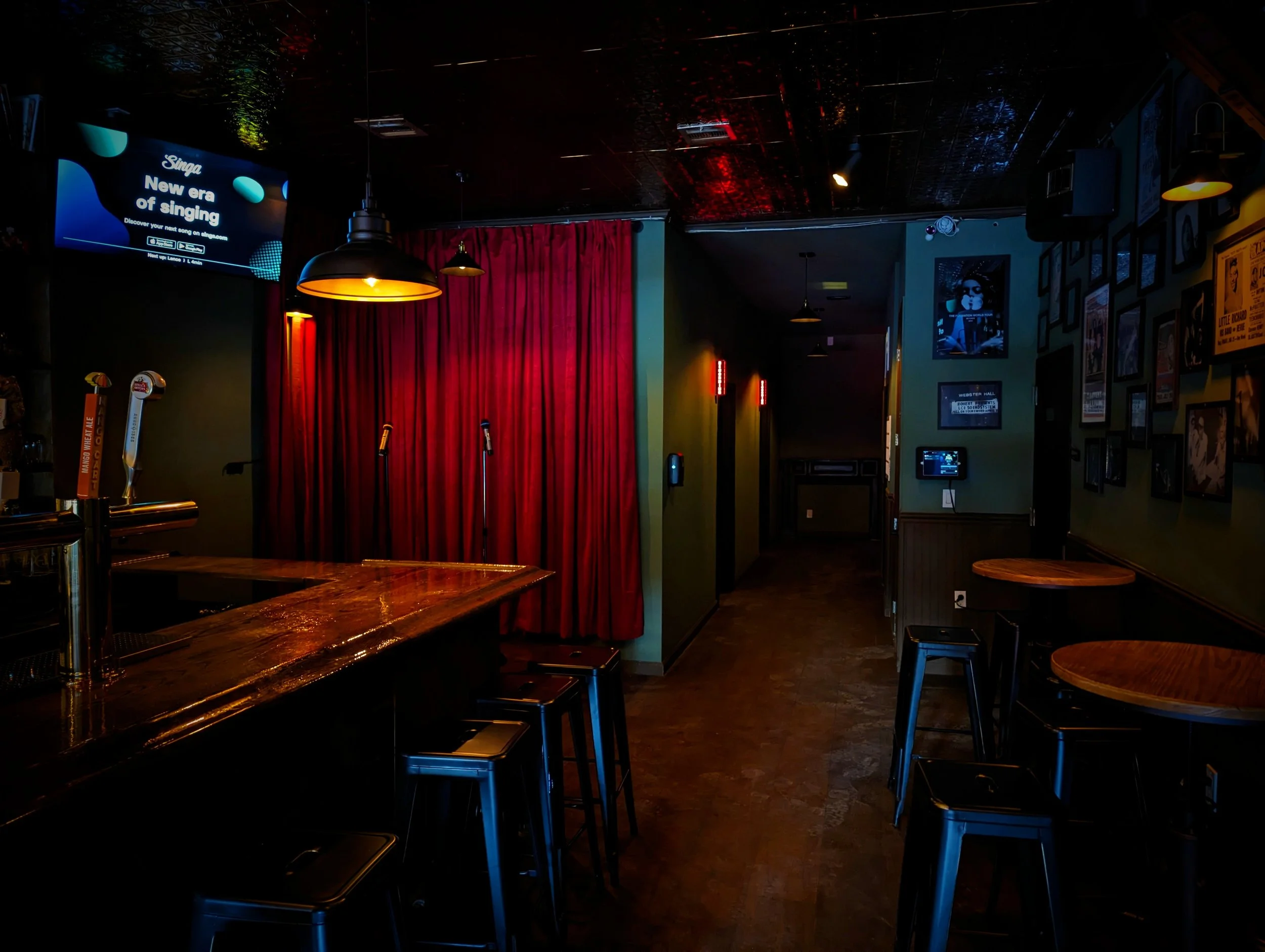 Dimly lit bar with a wooden counter, barstools, a red curtain, and framed pictures on the wall. TV screen shows advertising for a singing app, and there are microphone stands on the stage area.