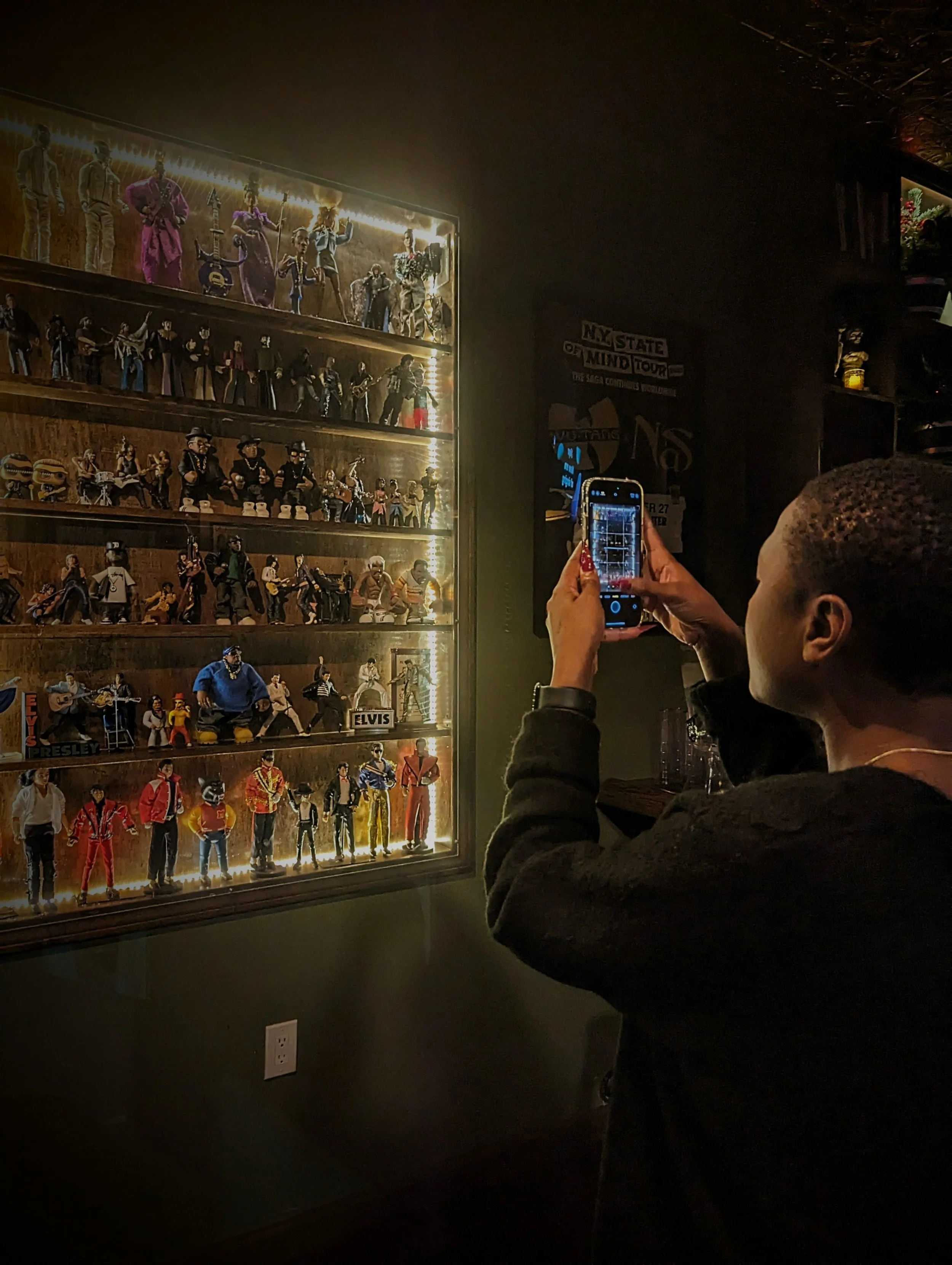 A person taking a photo of a display case filled with miniature figures of famous musicians, including Elvis Presley, The Beatles, and others, in a dimly lit room.