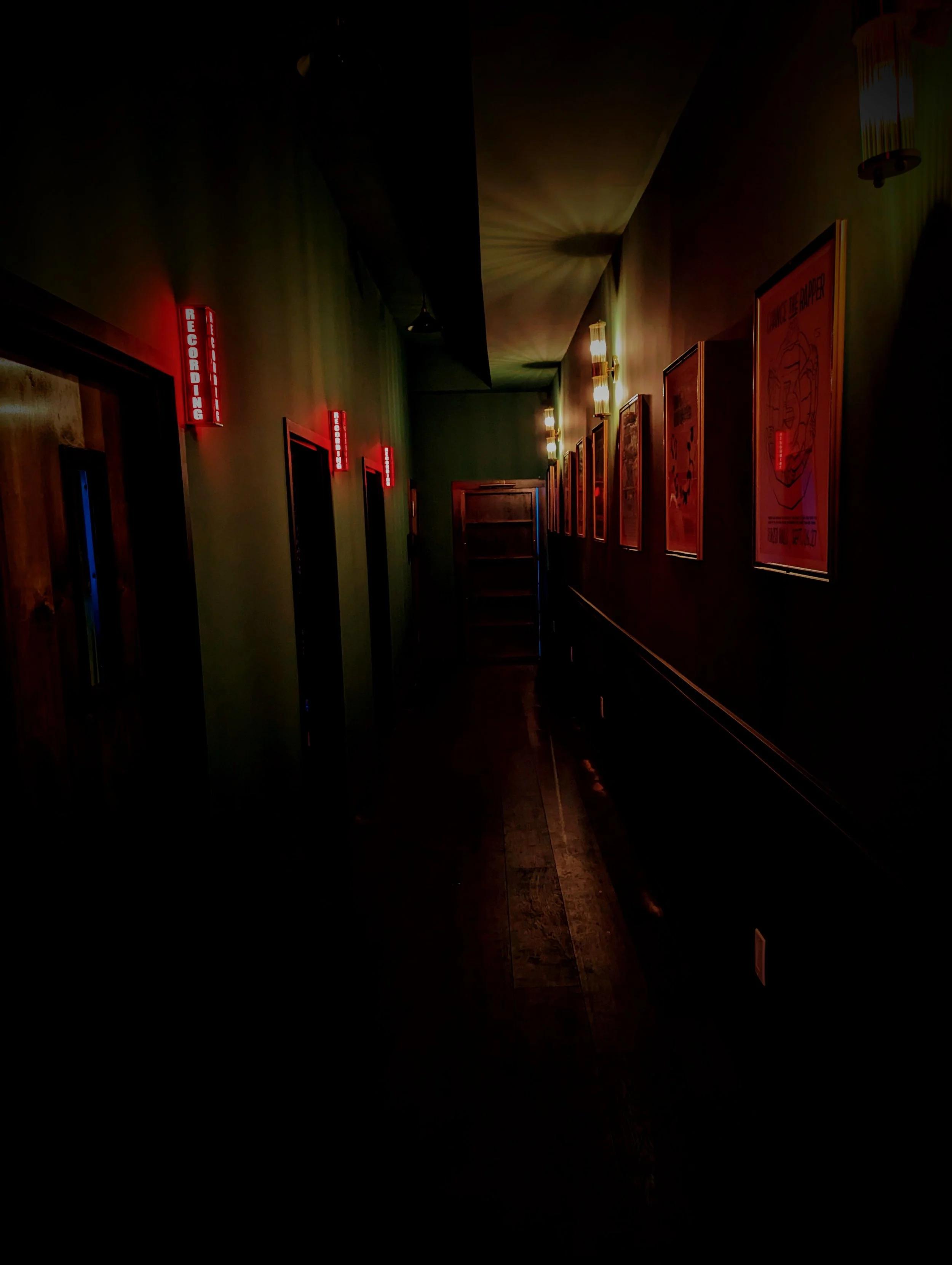 Dimly lit corridor with framed posters on the right wall and illuminated signs on the left wall.