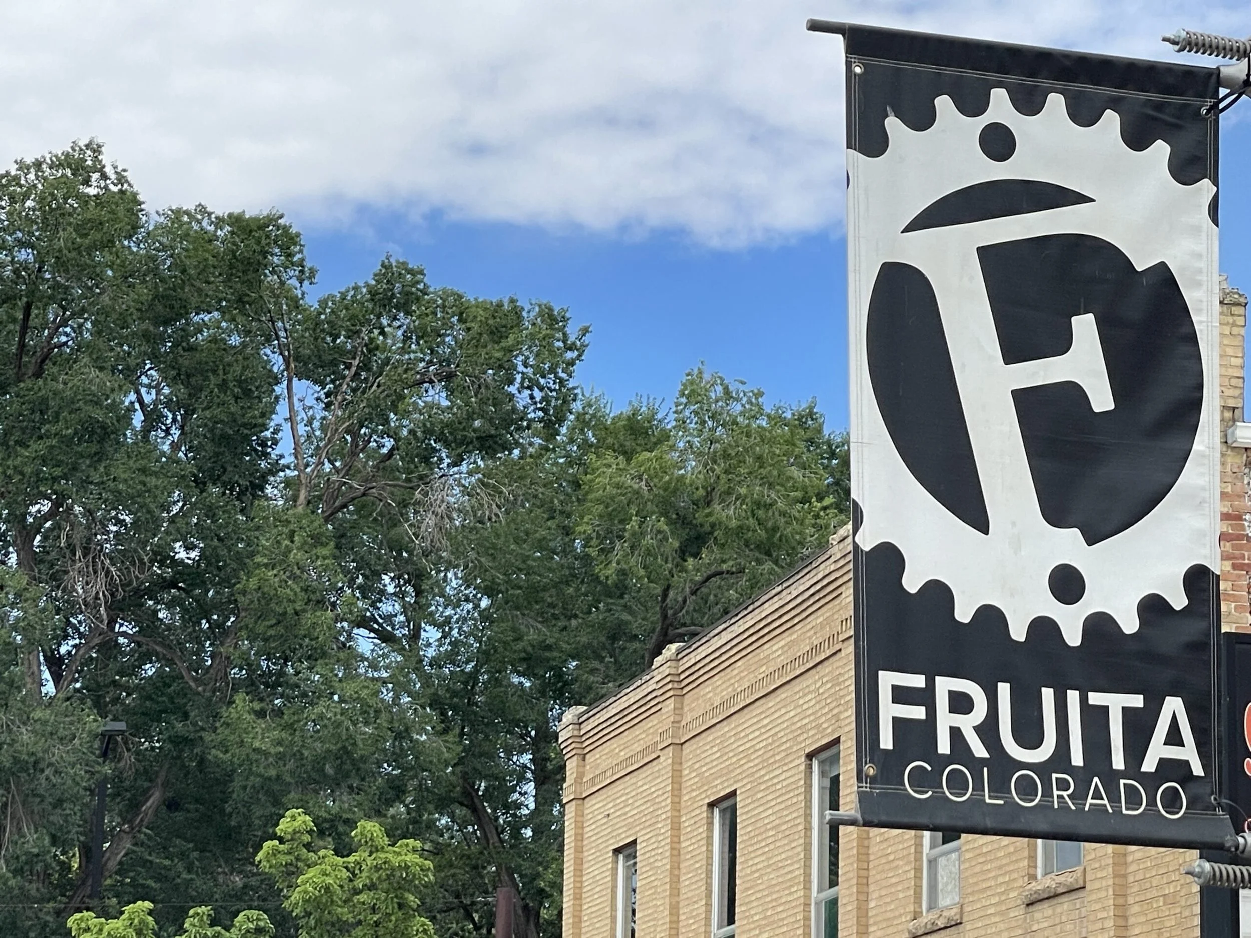 A black and white sign with a gear or cogs design and the text "FRUTA COLORADO" hanging outside a brick building surrounded by trees with green foliage and a cloudy sky in the background.
