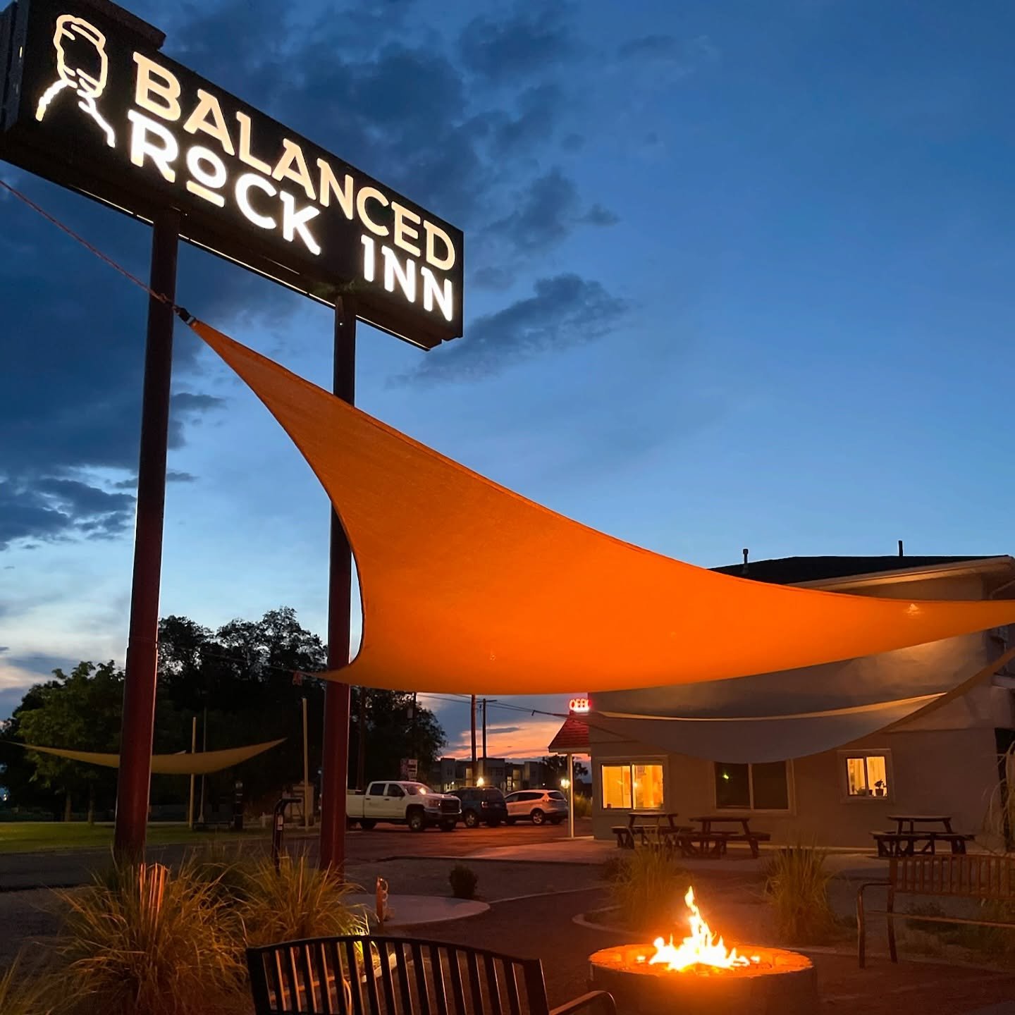 Outdoor evening scene at Balanced Rock Inn with a lit fire pit, canopy shade, building with warm lights, and parked cars against a darkening sky.