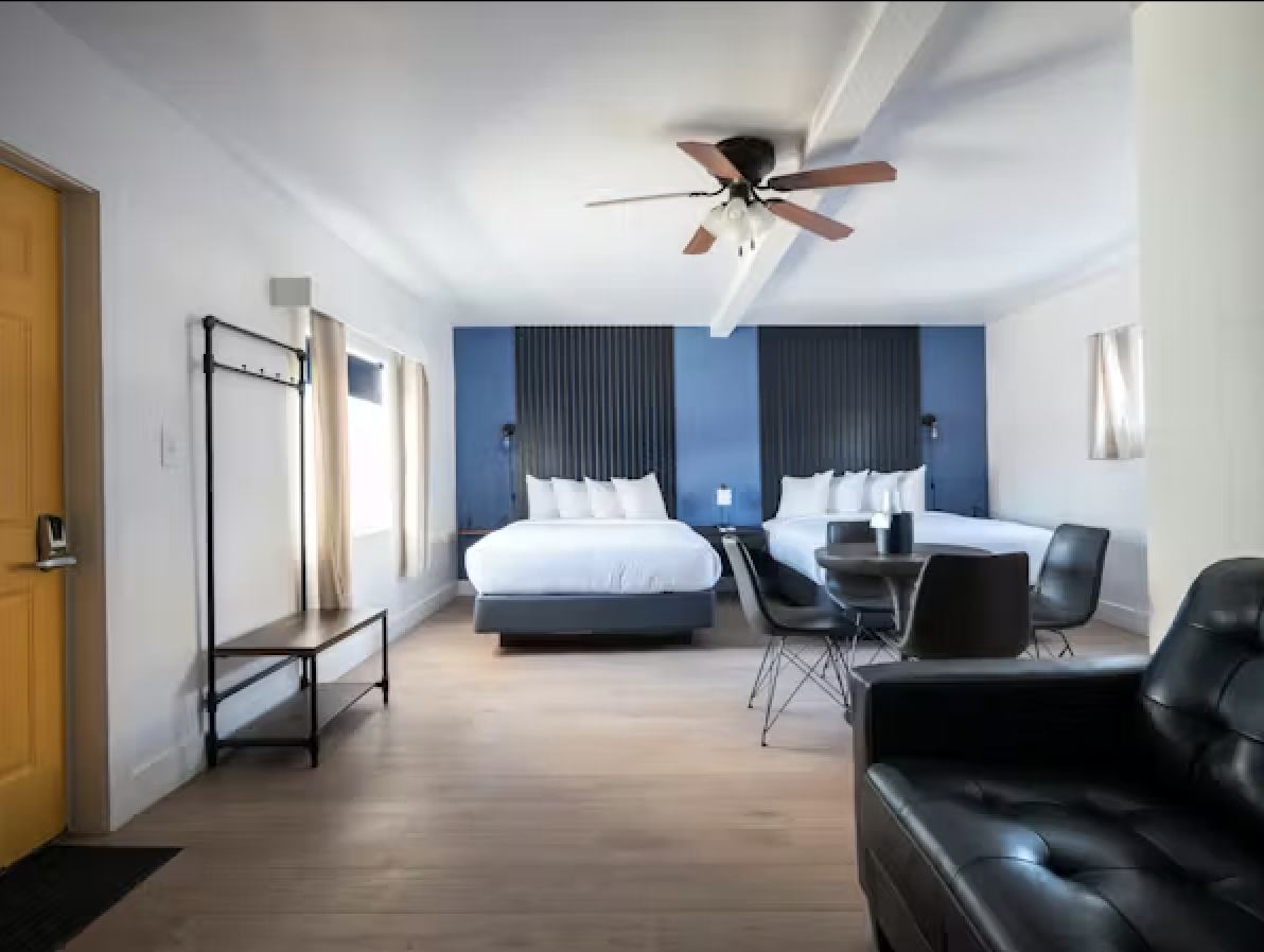 A hotel room with two queen beds, a black armchair, a round dining table with four black chairs, a ceiling fan, and windows with beige curtains.