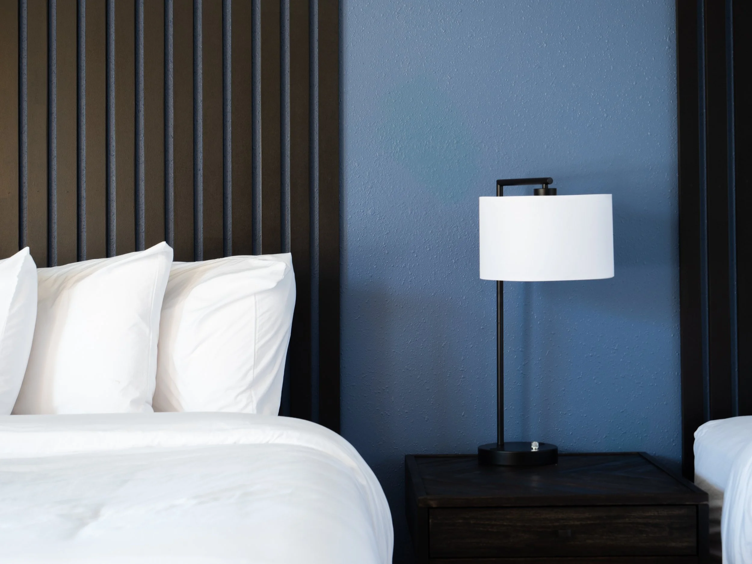 Close-up of a modern bedside table with a black metal lamp on a dark wooden surface, next to a bed with white pillows and sheets, against a textured blue wall with vertical black accents.