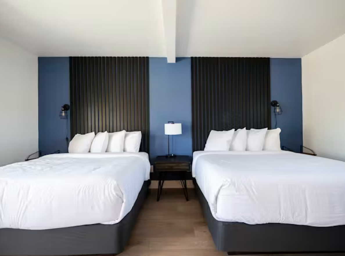 A hotel room with two double beds, white bedding, pillows, a dark wood nightstand with a lamp between the beds, and black wall-mounted reading lamps.
