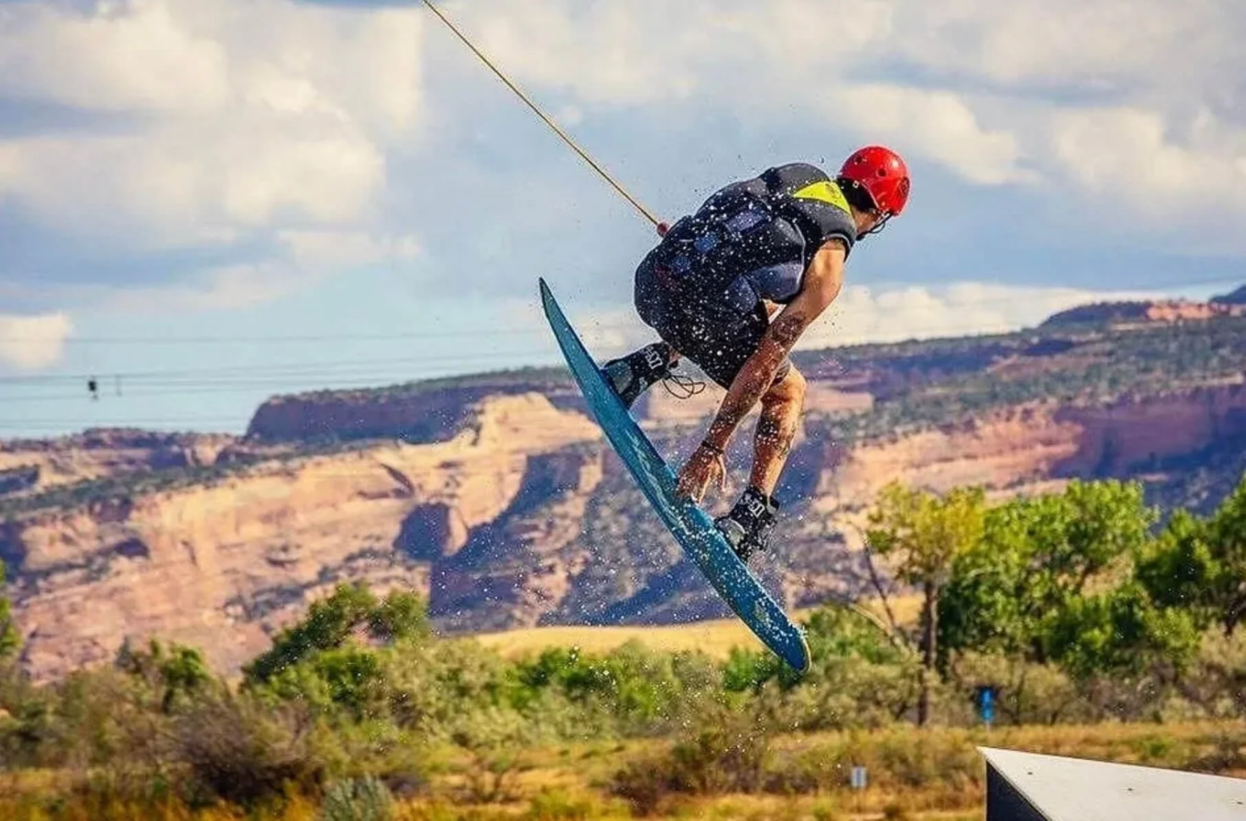 A person wearing a red helmet and black athletic clothing is mid-air on a wakeboard, performing a jump with a scenic canyon landscape in the background.
