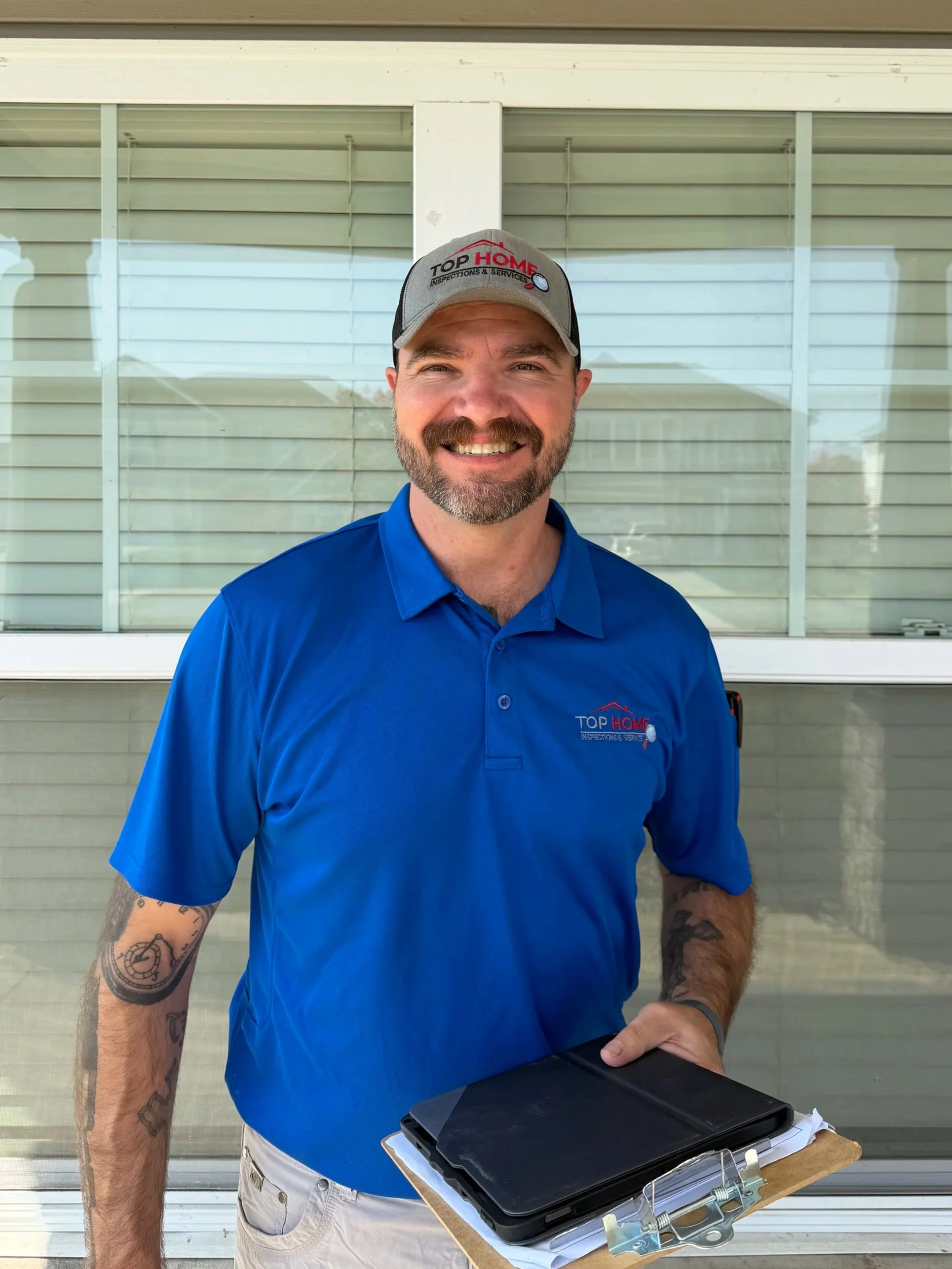 A smiling man wearing a gray cap and blue polo shirt with a logo, standing outside near a window with horizontal blinds, with a clipboard and tablet on a stand in front of him.