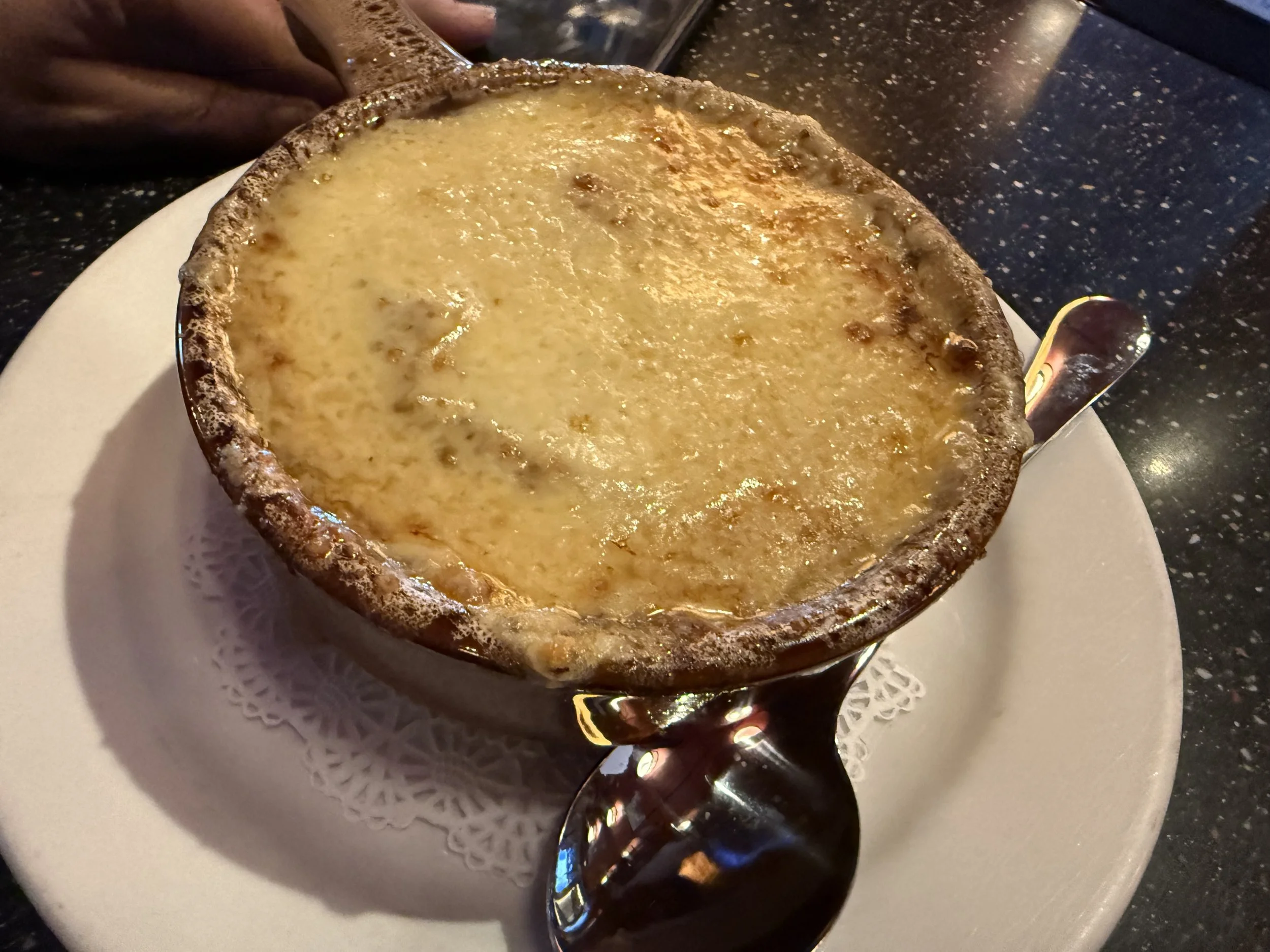 A small bowl of melted cheese dip with bubbles on the surface, resting on a white doily on a white plate, with a spoon beside it.