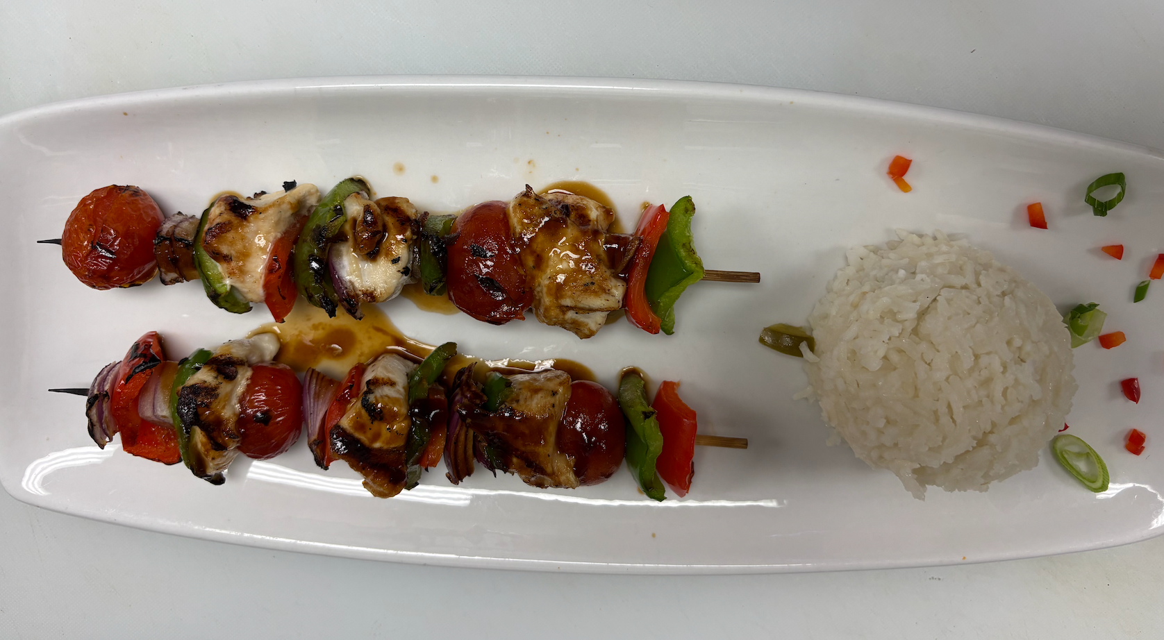 Two vegetable skewers with cherry tomatoes, bell peppers, onions, and chicken, served with a side of white rice, garnished with chopped green onions and small red and green pepper pieces.