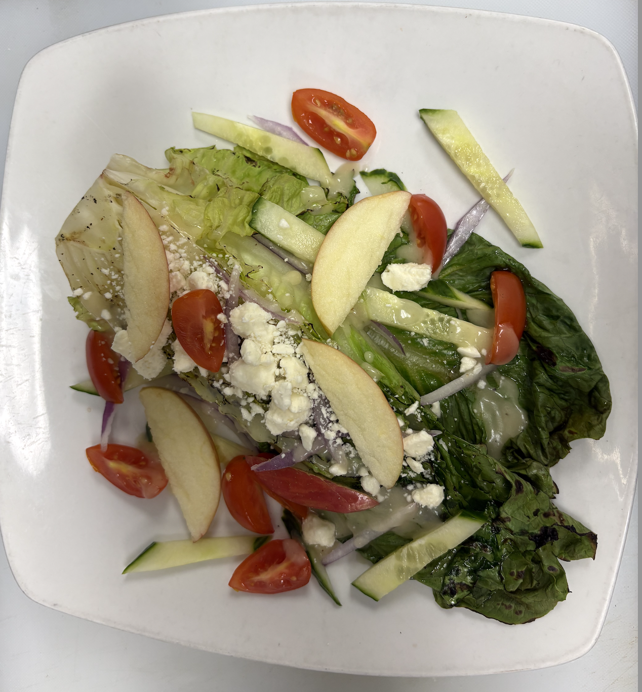 Mixed green salad with cherry tomatoes, sliced cucumbers, sliced apples, red onions, crumbled cheese, and a creamy dressing on a white square plate.