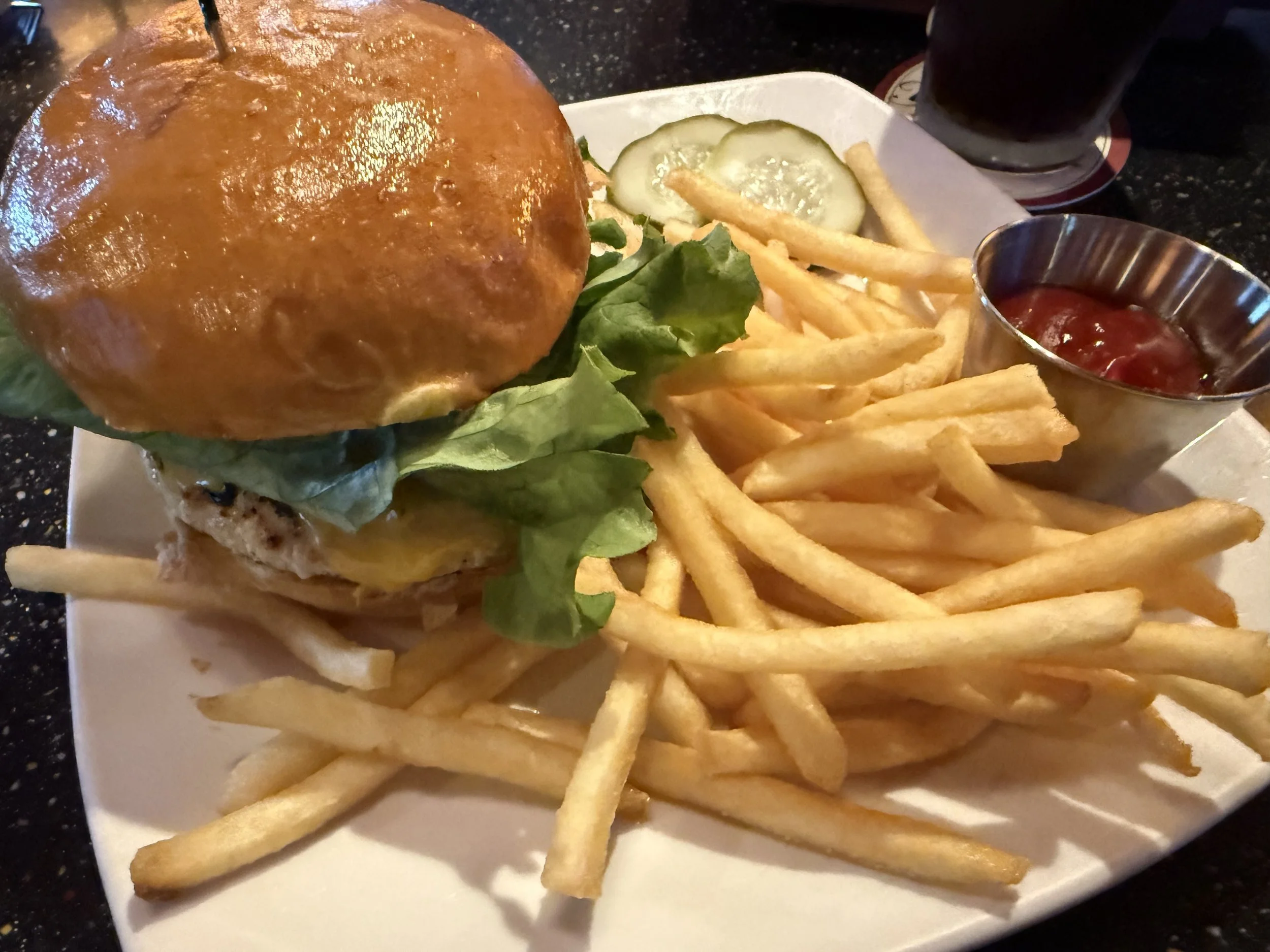 A plate with a cheeseburger, lettuce, sliced pickles, French fries, ketchup, and a glass of dark soda.