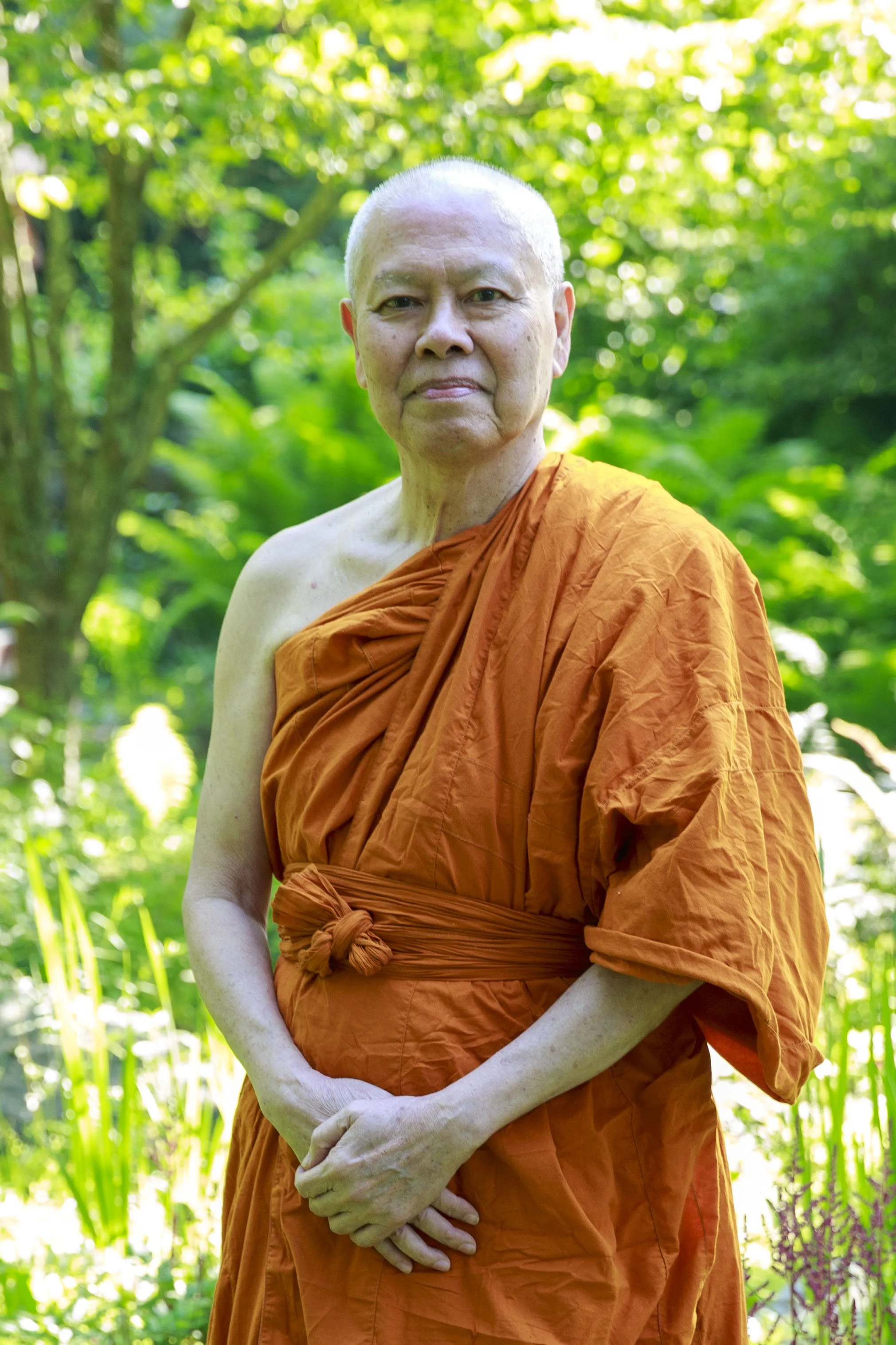A Buddhist monk wearing an orange robe standing outdoors in a lush, green forest.