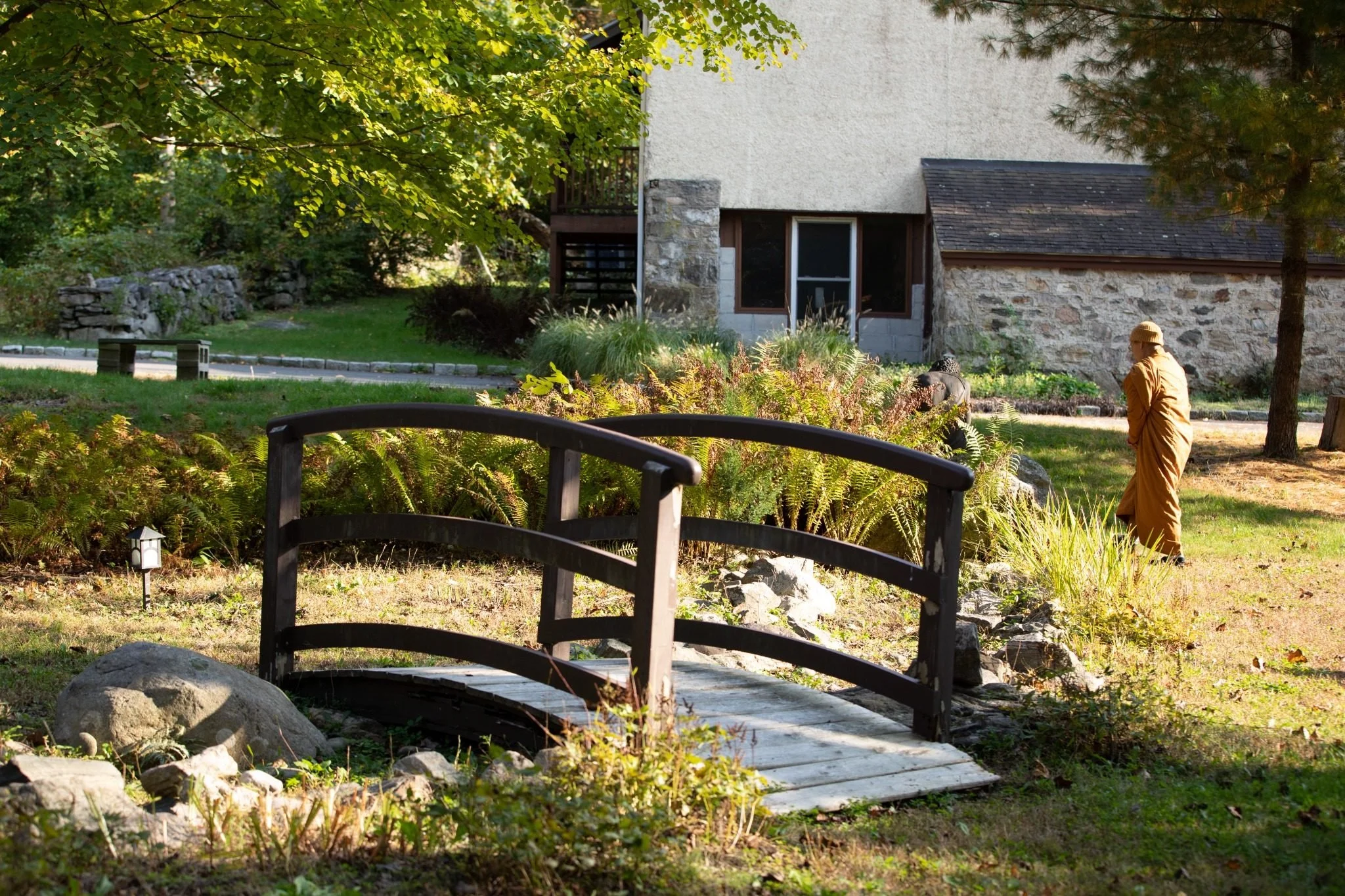 A landscaped garden with a small wooden bridge over rocks, surrounded by lush greenery, a stone and wood building in the background, and a person in a long coat and hat walking along the garden path.
