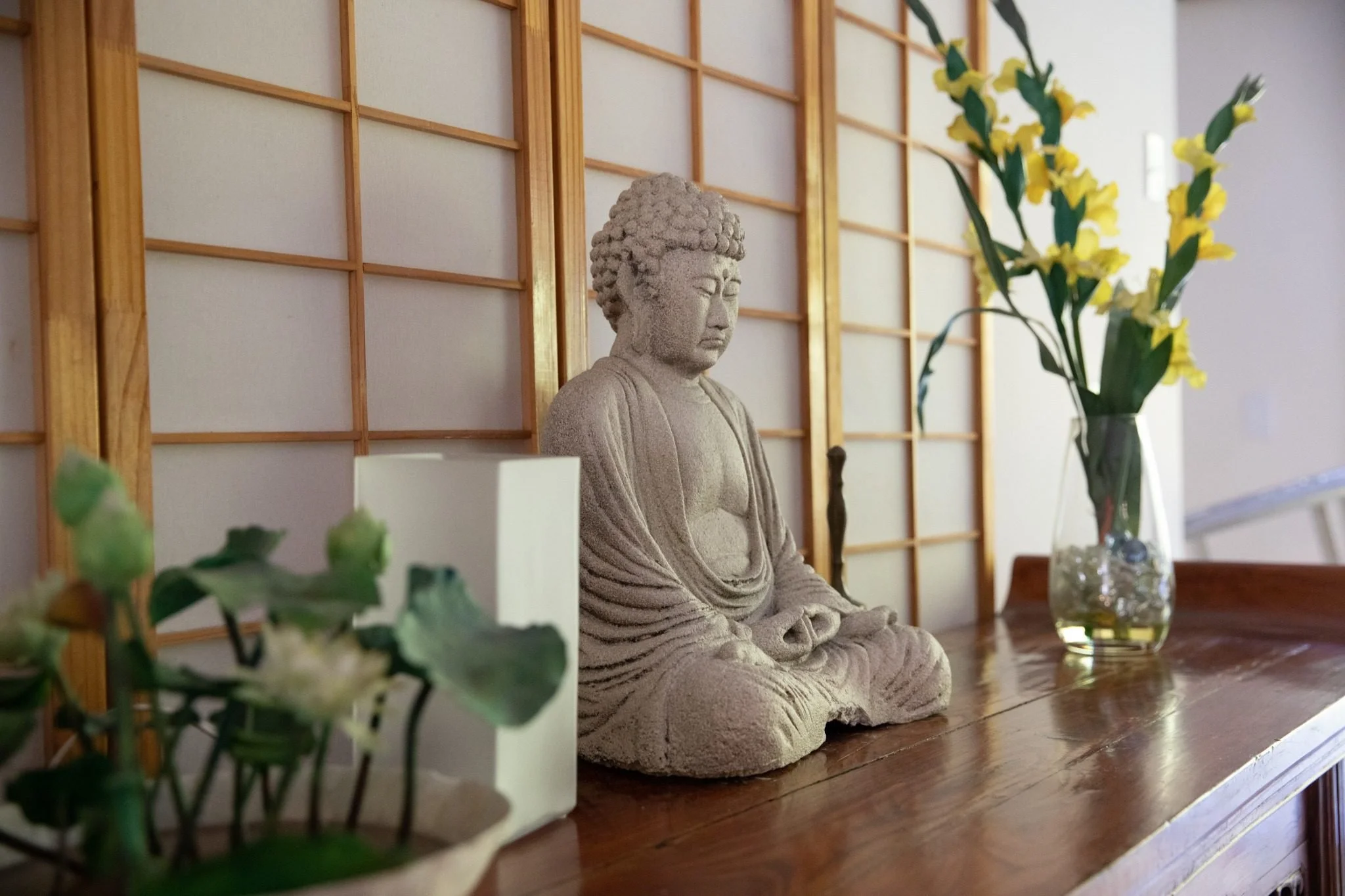 A stone statue of Buddha sitting cross-legged on a wooden surface, with a background of a wooden lattice wall and a vase of yellow flowers.