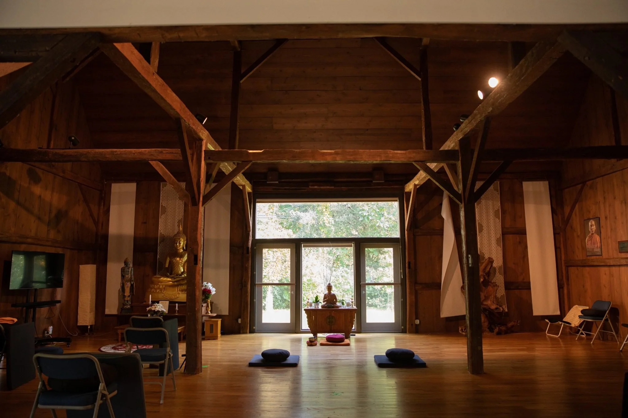 A spacious wooden meditation room with a central altar featuring a Buddha statue, cushions on the floor, and chairs around the room, illuminated by natural light through large windows.